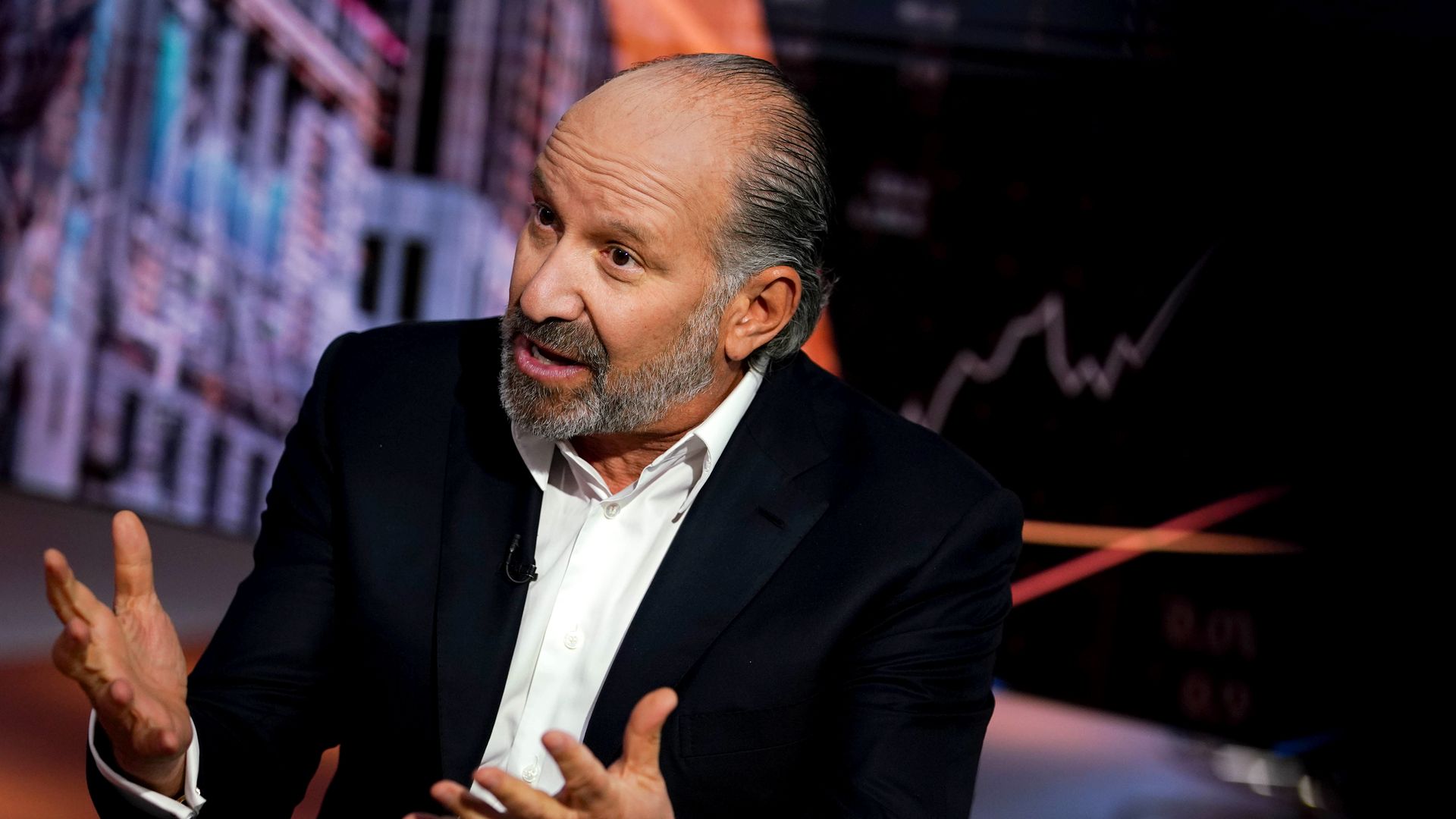 A bald person in a suit jacket and white shirt gestures with their hands