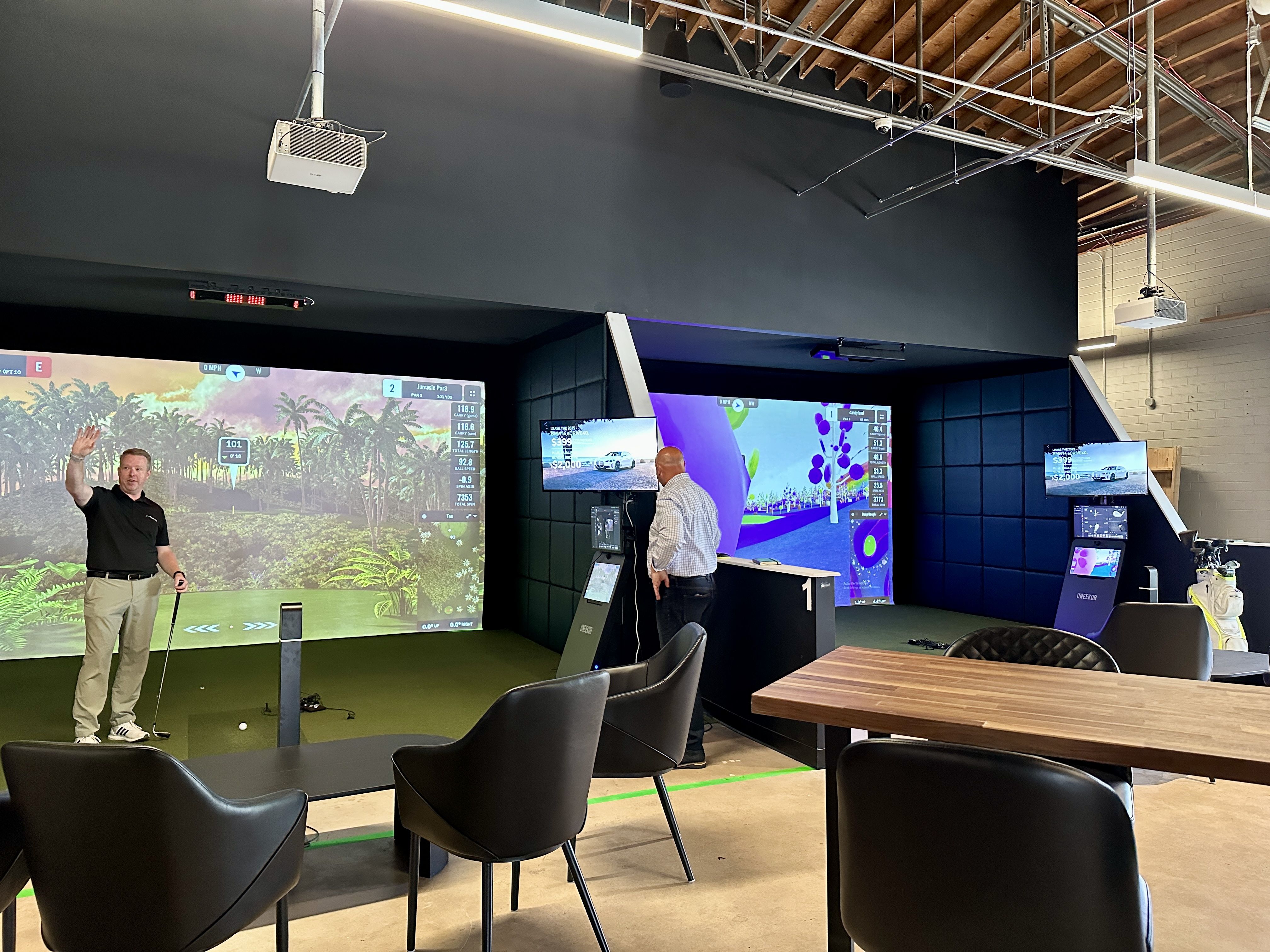 Indoor golf simulator club with two men, one holding golf club in black shirt and khaki pants, the other in checkered shirt, multiple large screens showing golf courses, black chairs, and wooden table.