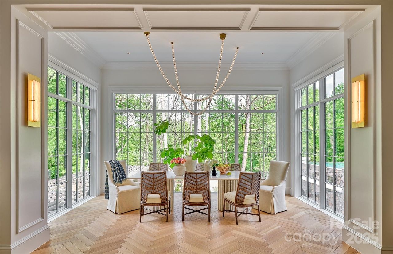 Bright dining room with large windows showing green trees outside, light wood herringbone floor, modern woven chairs, white armchairs, and a chandelier with hanging beads.