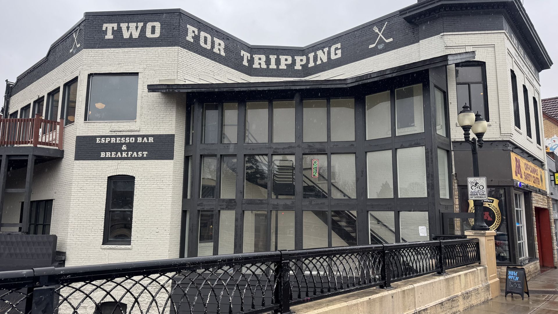 Corner brick building with large glass storefronts. The facade reads "TWO FOR TRIPPING" across the top; a sign says "ESPRESSO BAR & BREAKFAST". A railing and street lamps line the sidewalk.