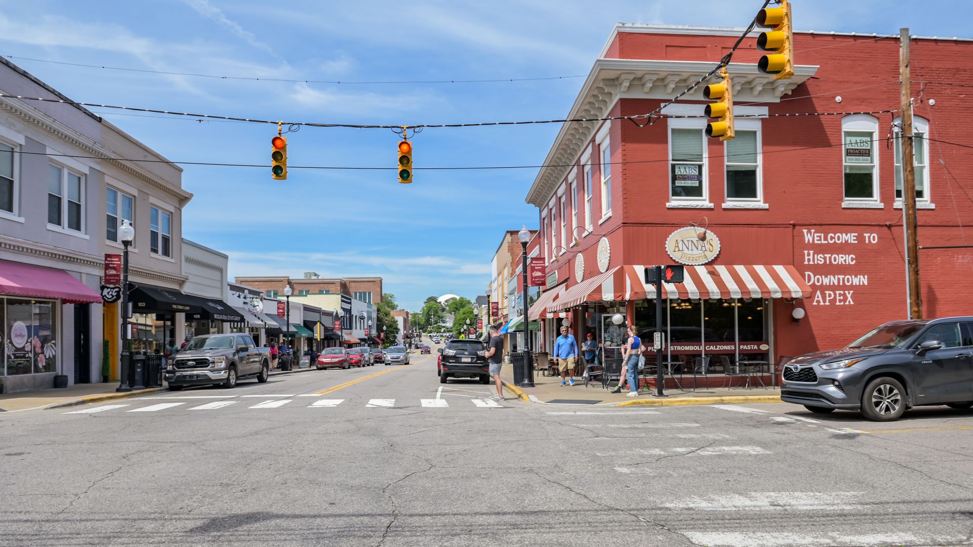 Main street of historic downtown Apex on a sunny day, showing red brick buildings, a red traffic light, Anna's Pizzeria with red-striped awning, parked cars, and pedestrians crossing.