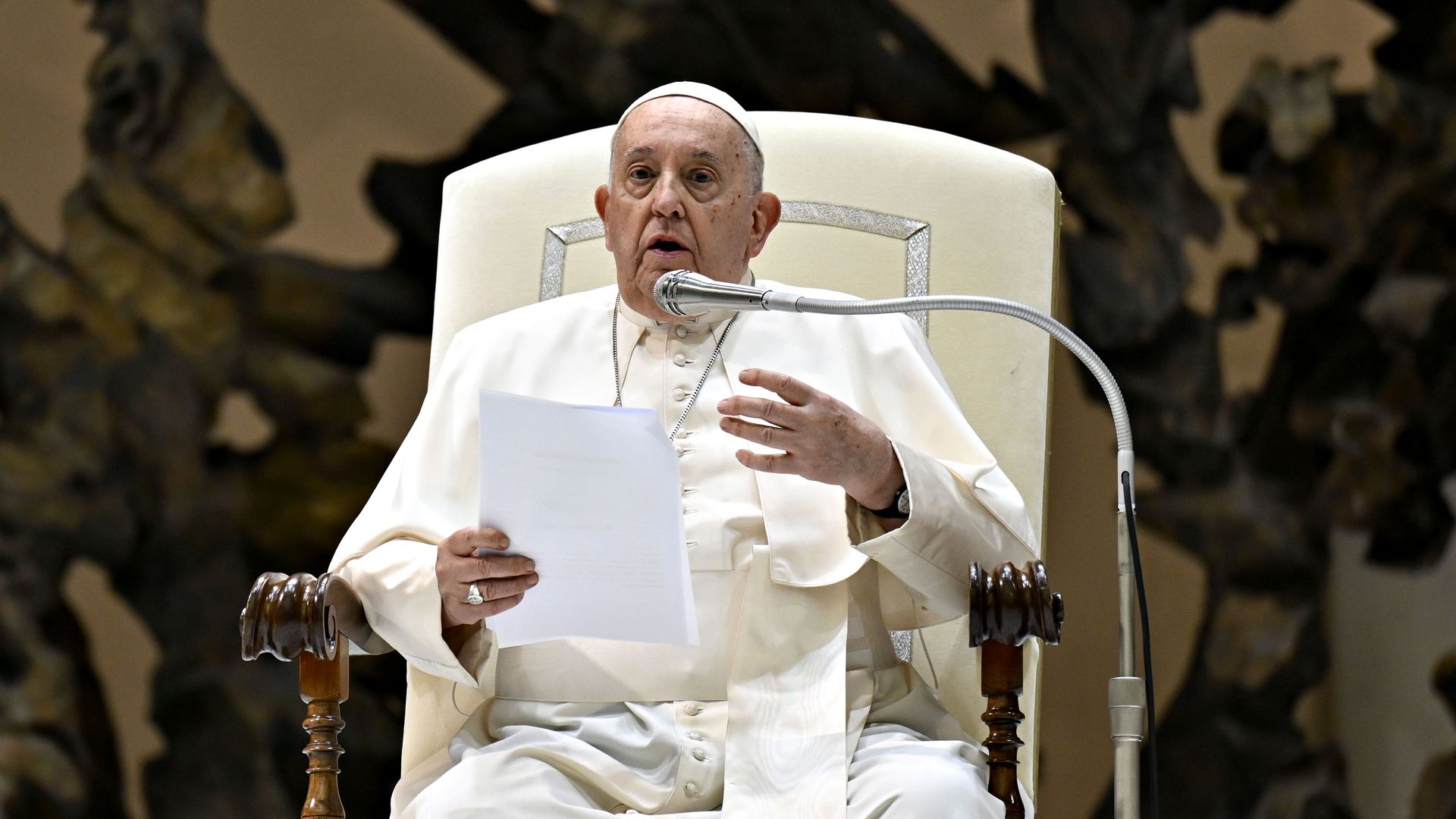 Pope Francis sits and speaks into a microphone. He holds a paper in one hand. 