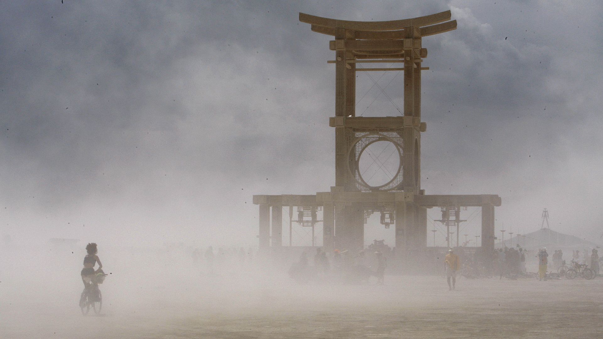 A dusty scene with a large wooden structure featuring a circular design in the center, blurred figures walking and a person riding a bicycle in the foreground under cloudy skies.