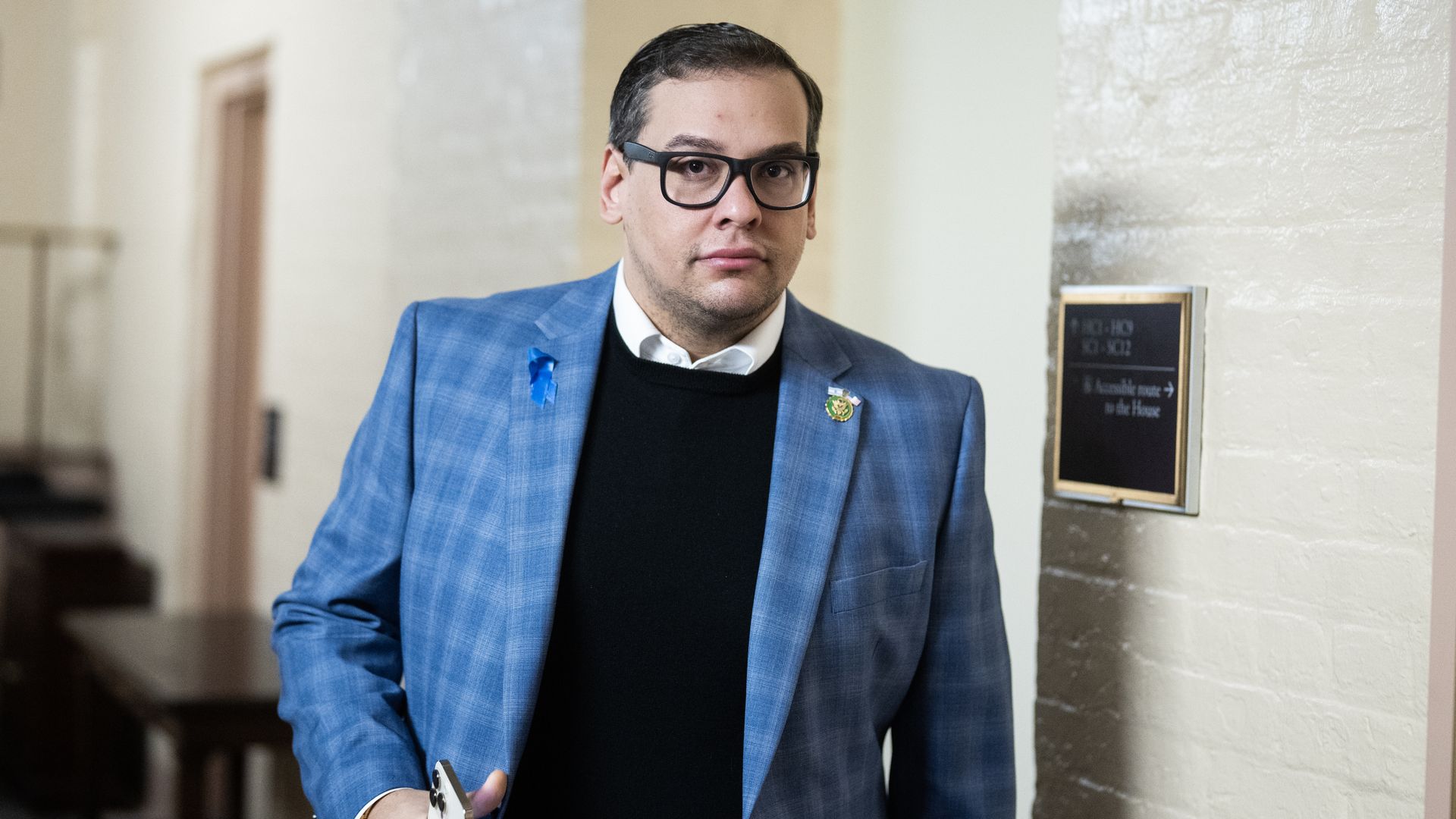 Former Rep. George Santos, wearing a blue jacket, dark blue sweater and glasses, walks in a white brick hallway.