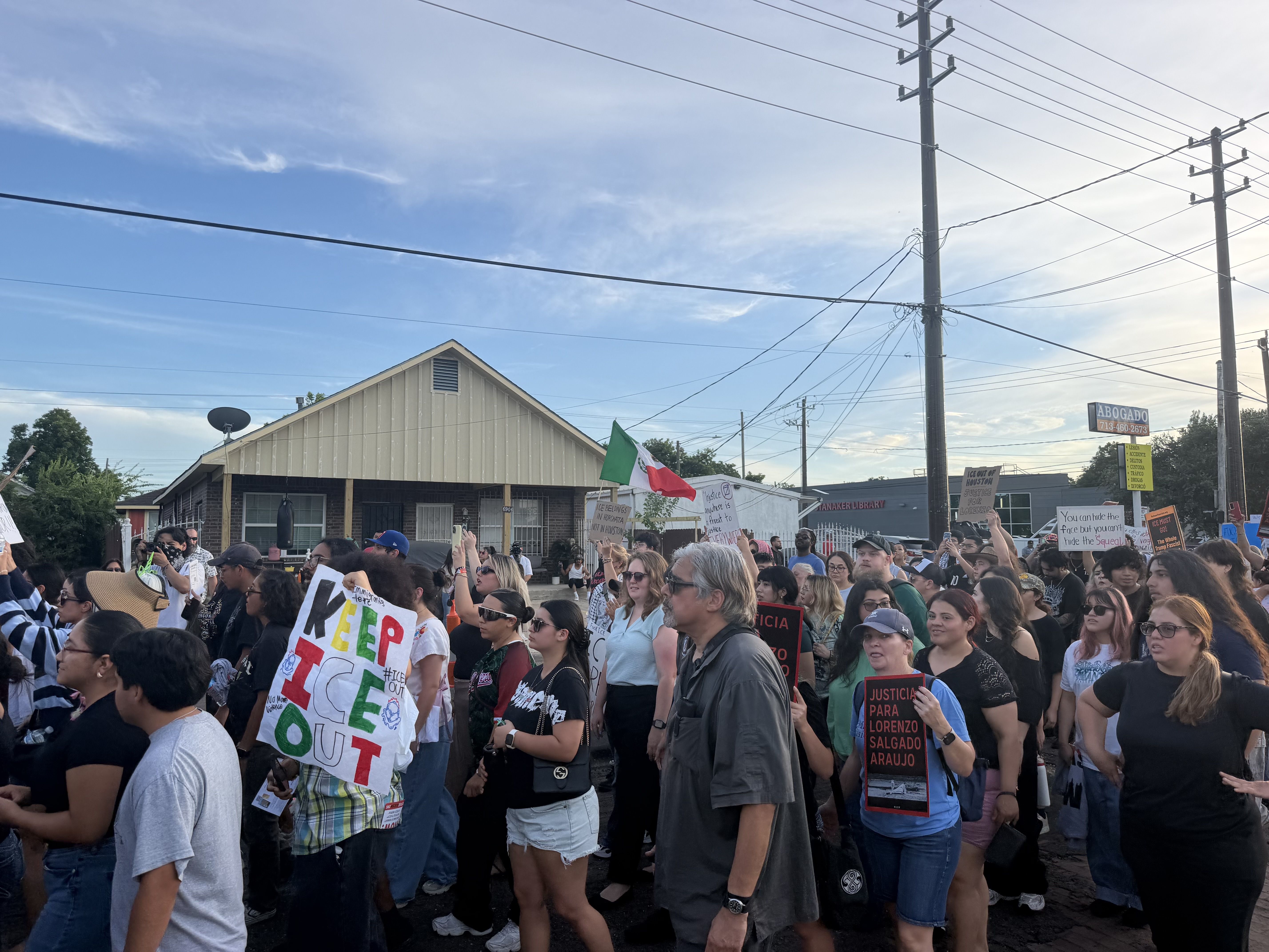 Large crowd of diverse protesters fills a city street, many holding signs. Power lines and poles cross the sky; a Mexican flag is visible with buildings and an "ABOGADO" sign nearby.