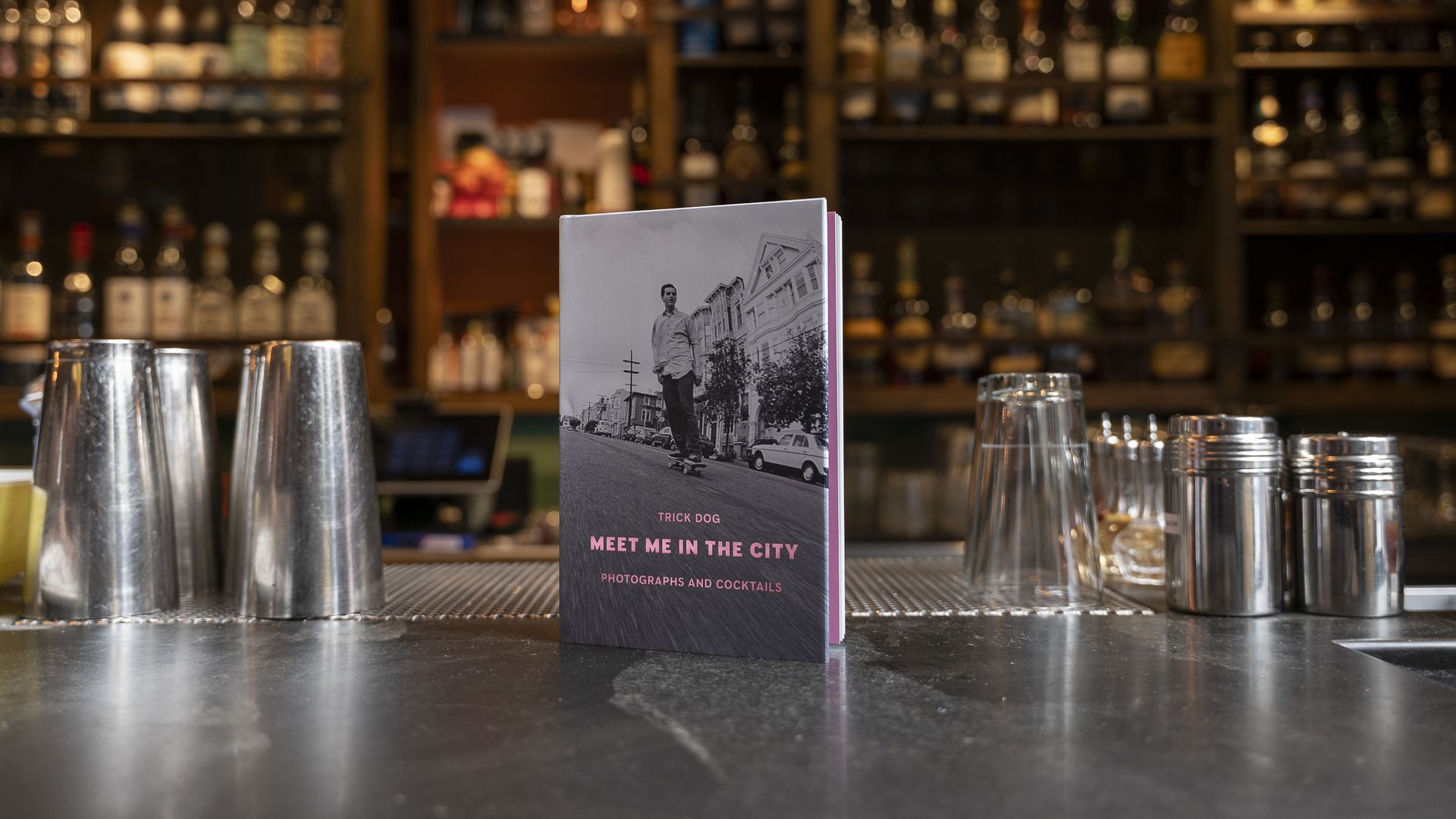 Menu titled "Meet Me in the City" by Trick Dog with image of man skateboarding, placed on bar counter surrounded by metal shakers and glasses, bottles blurred in background.