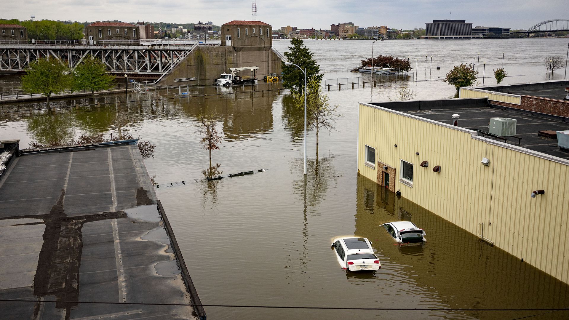 In this image, two cars float in the flood water between two buildings. This picture is a birds eye view of flood waters in a city.