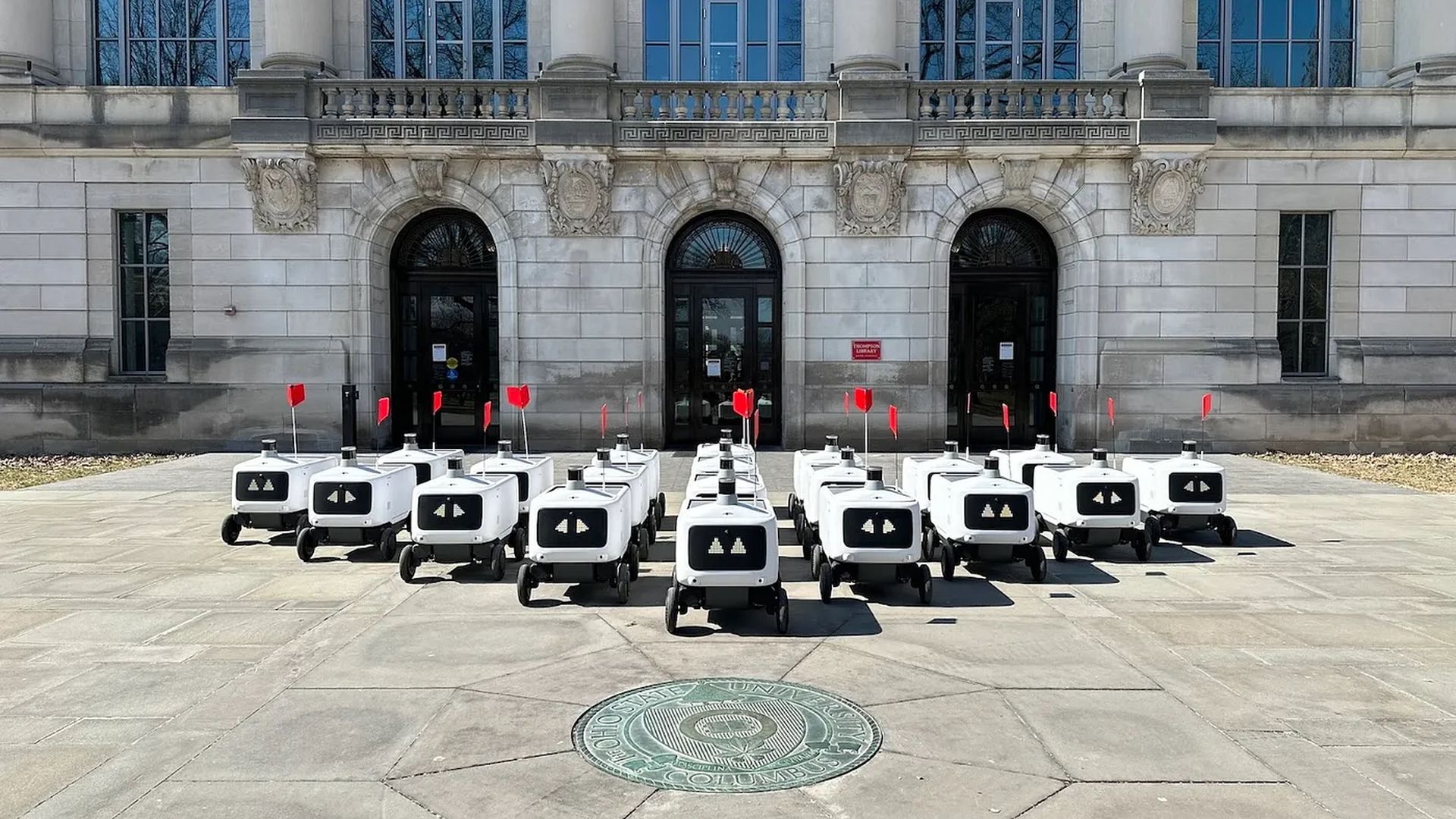 Rows of white food robots topped with red flags and bearing screen faces with digital eyes on Ohio State's campuses