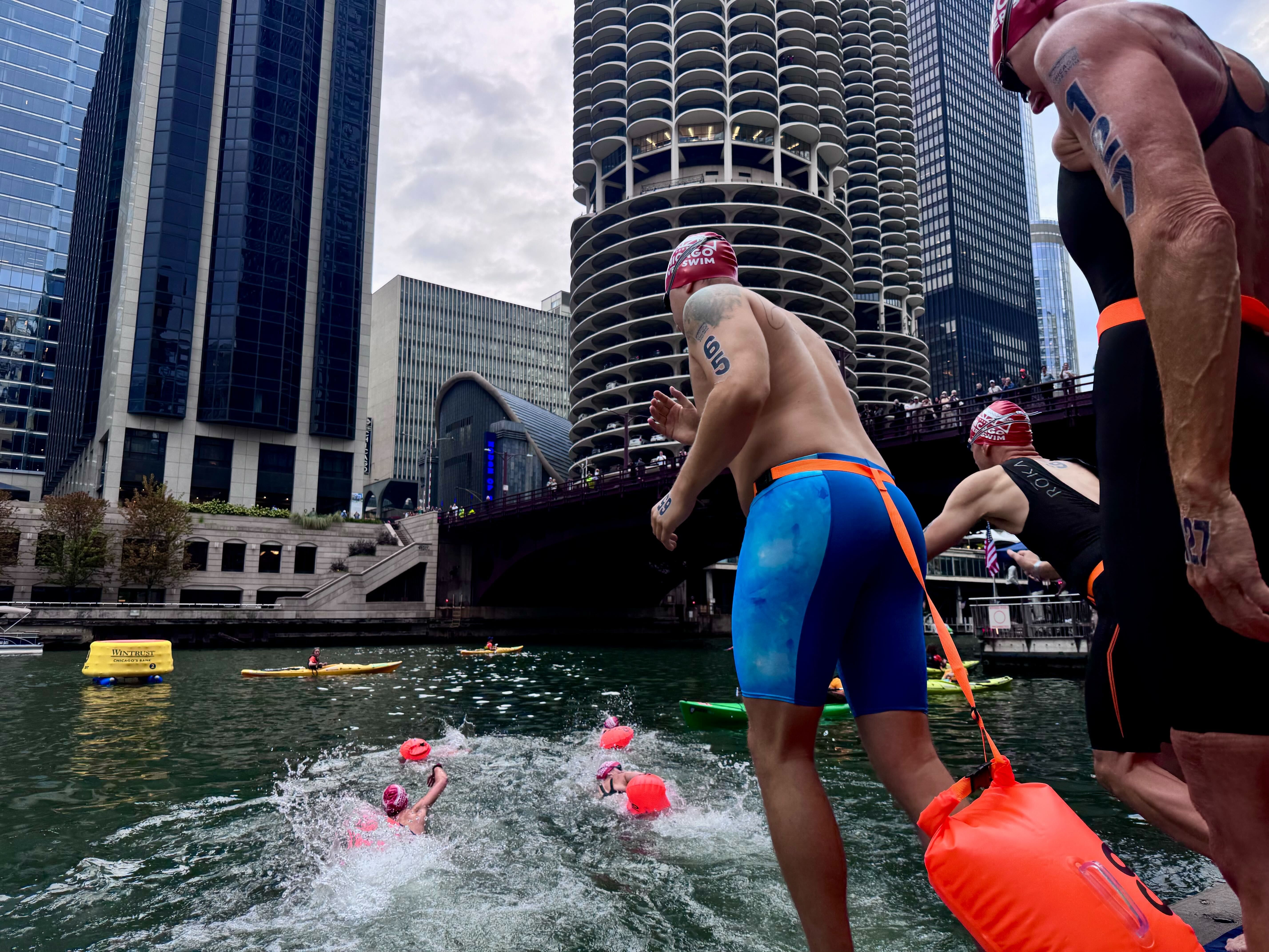 Swimmers in red caps and blue or black swimwear diving into a river in a city with tall buildings and unique round balconies, some spectators watching from a bridge above.