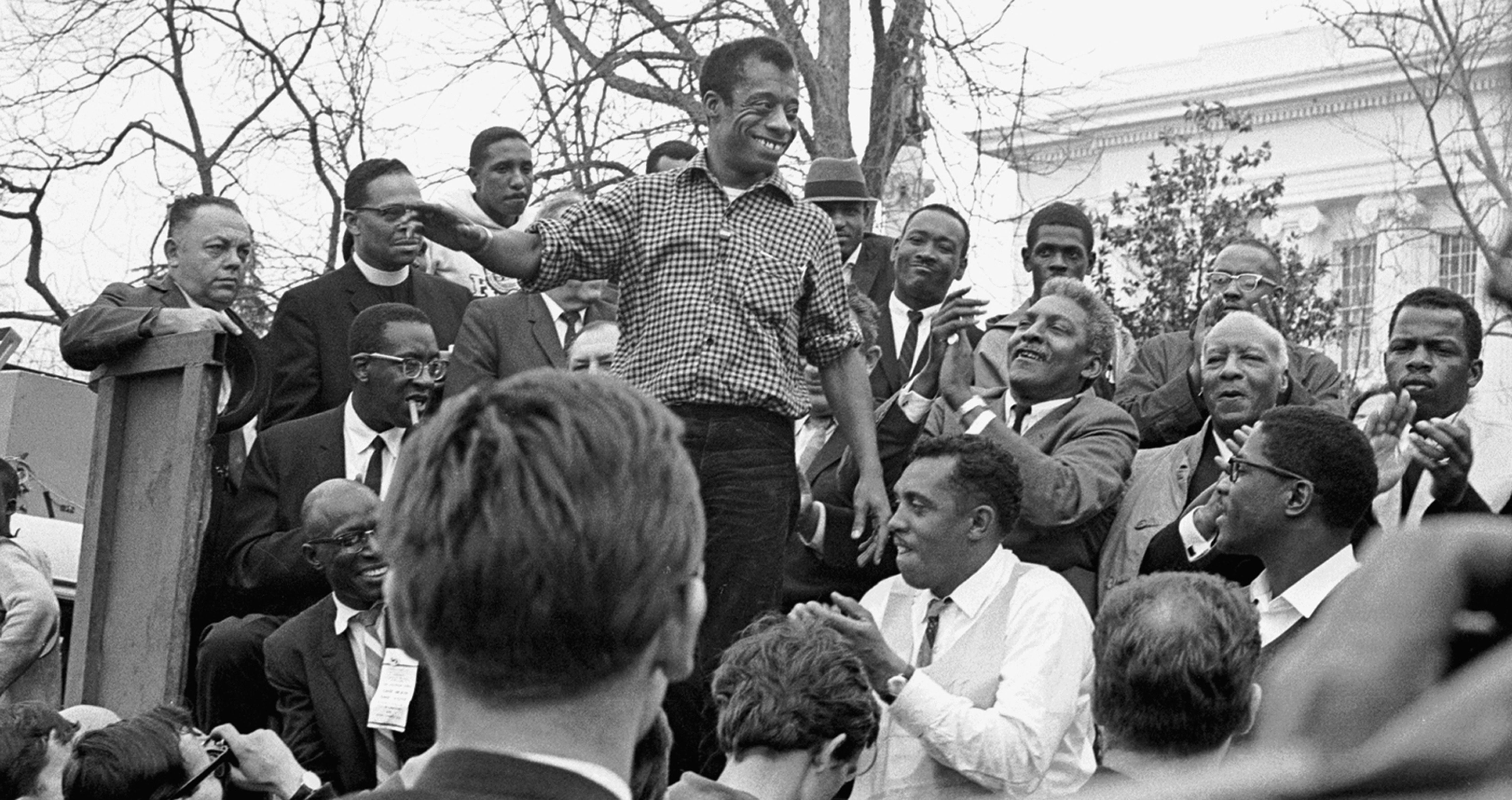 James Baldwin stands, smiling, in a button down shirt. He is looking around at a crowd of people who are all looking at him