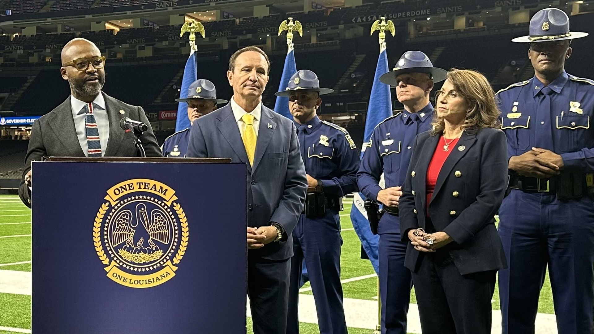 A man stands in front of a gathering of other people at a podium on the football field of the Superdome.