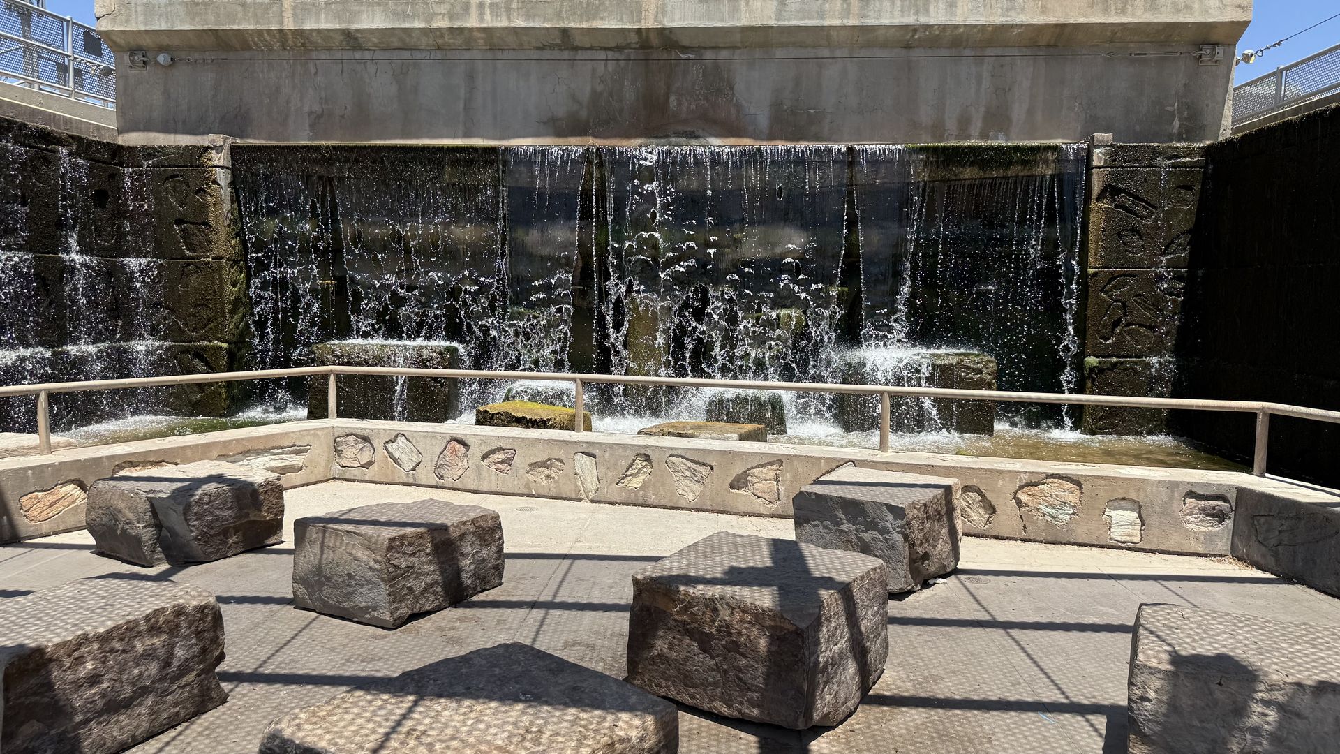 A cascading waterfall feature with several stone block seats with rough hewn sides, separated by a metal railing. 