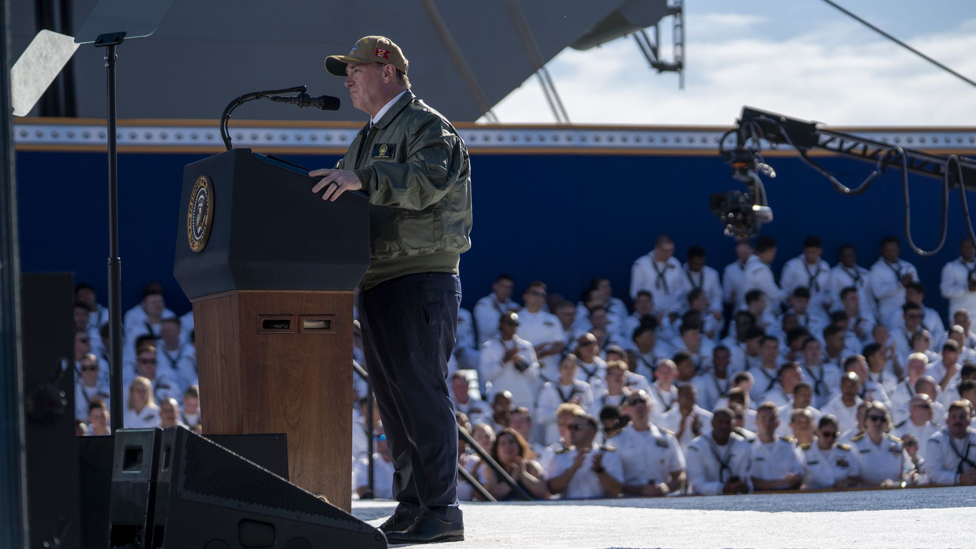 Man in military green jacket and cap speaking at a podium with the presidential seal, in front of seated navy personnel in white uniforms on ship deck under sunlight.