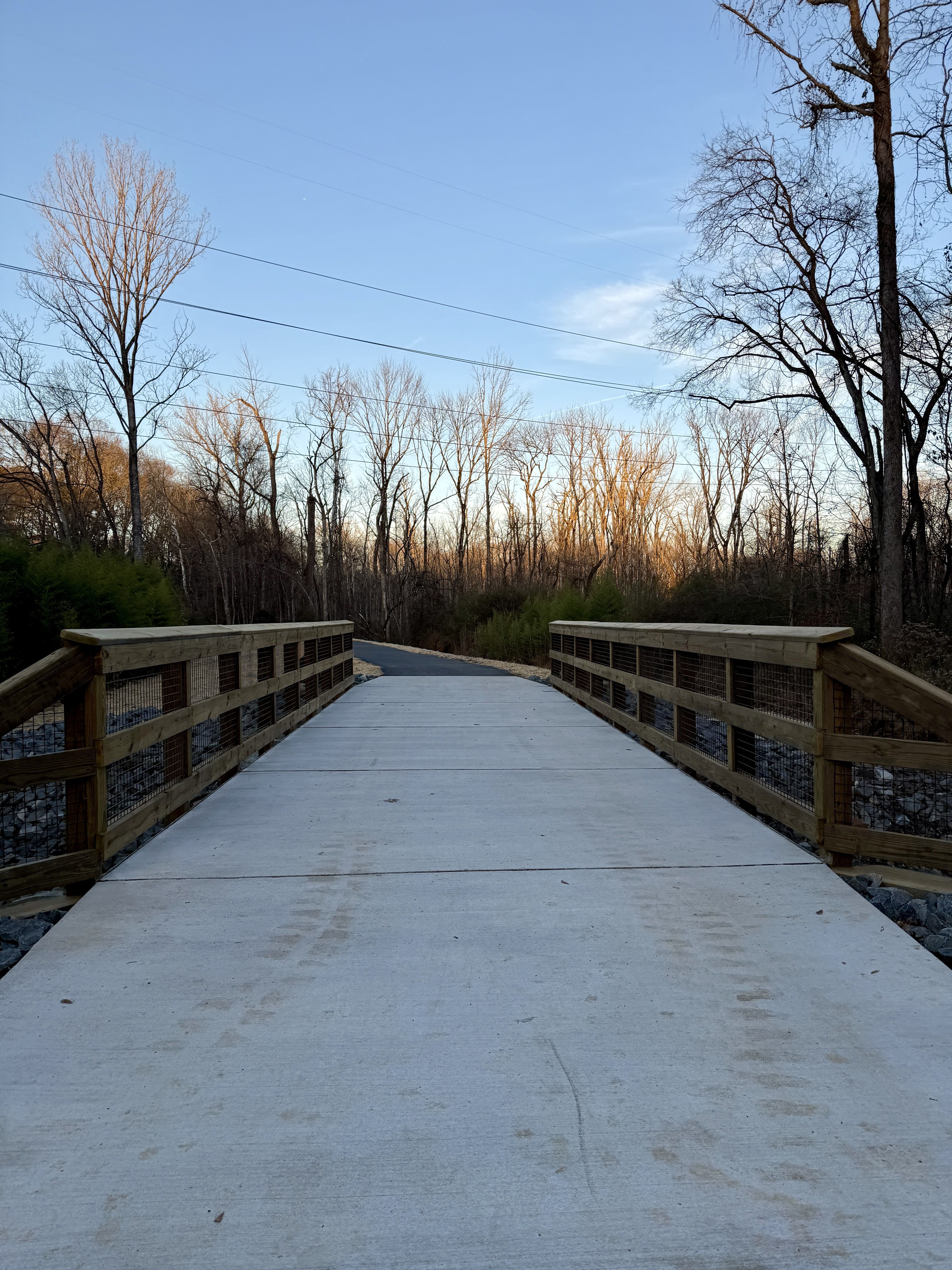 Concrete bridge with wooden railings over rocky ground, leading to a winding paved path through leafless trees at sunset under a clear blue sky.