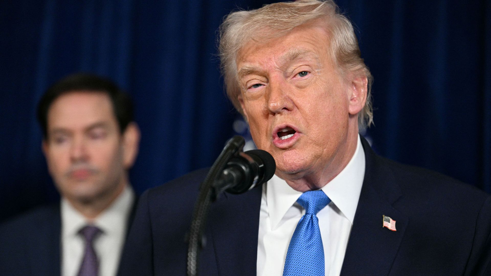 President Donald Trump at a podium speaking to reporters outdoors with Secretary of State Marco Rubio standing behind him.