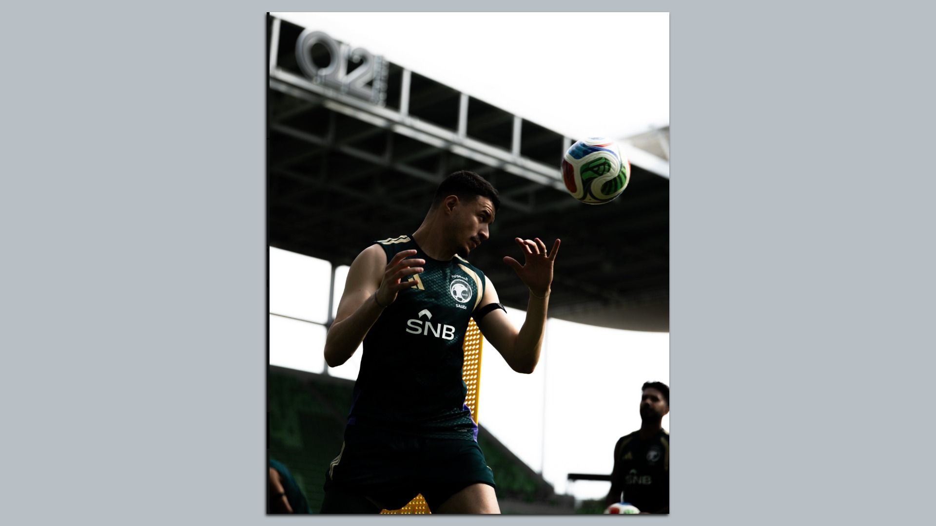 A male soccer player in a dark green SNB jersey, under stadium beams, watches as a colorful ball flies toward his head during a game.