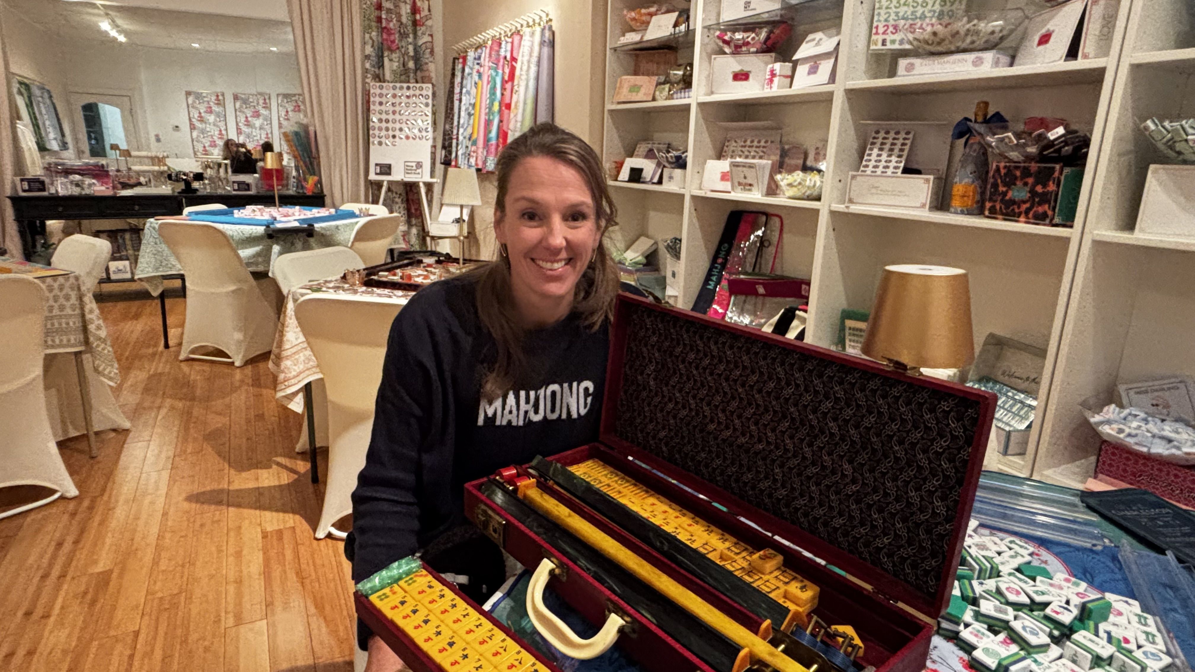 Smiling woman in a black "MAHJONG" shirt sits in a cozy room with white shelves and tables covered with cloths, holding an open mahjong set case with yellow and green tiles.