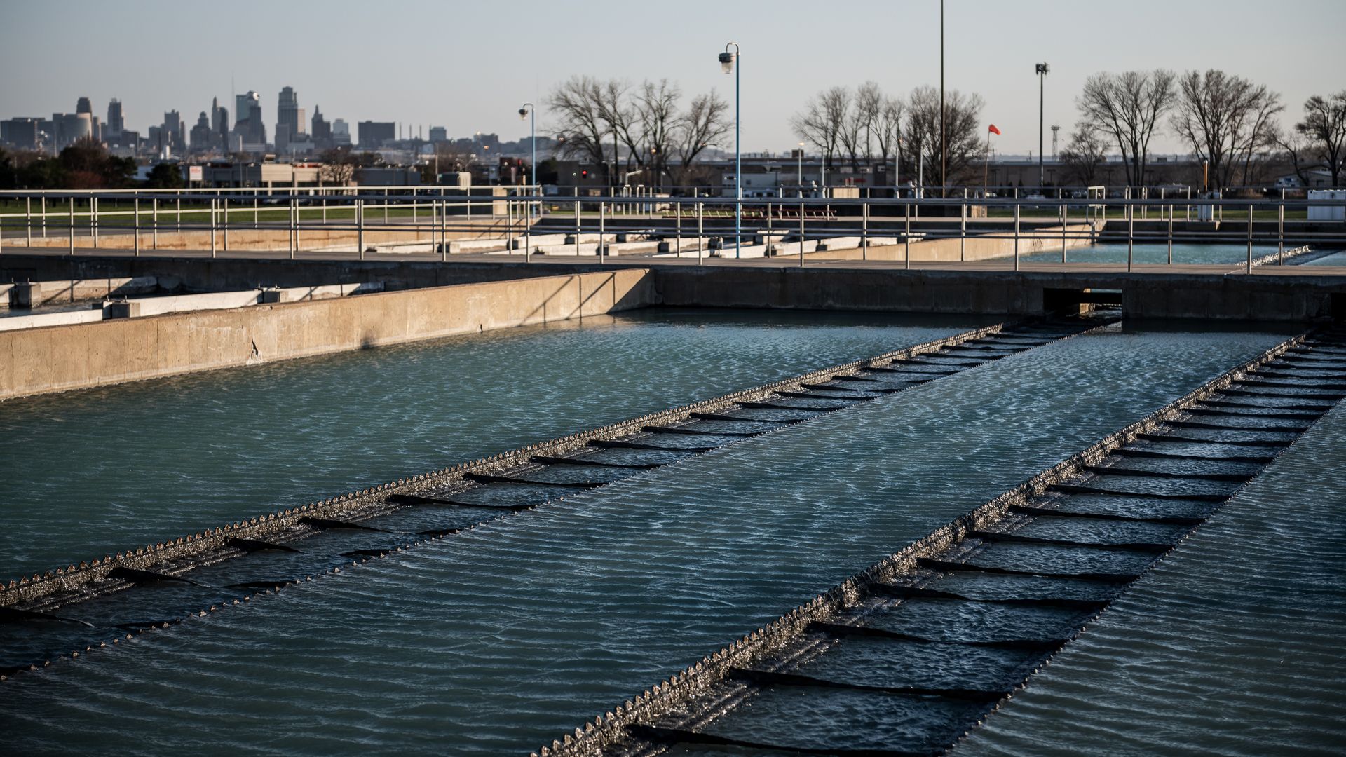 Outdoor water treatment facility with multiple concrete basins filled with blue water, metal railings, and a city skyline in the background under a clear sky.