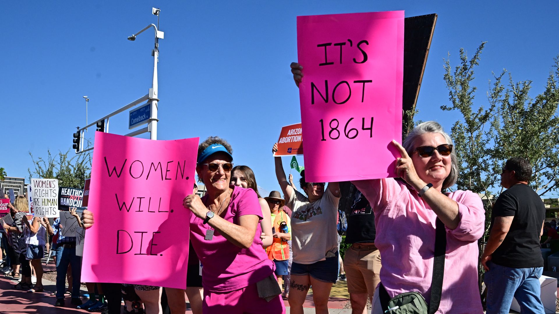 Women holding pink signs that say, "IT'S NOT 1864" and "WOMEN. WILL. DIE."
