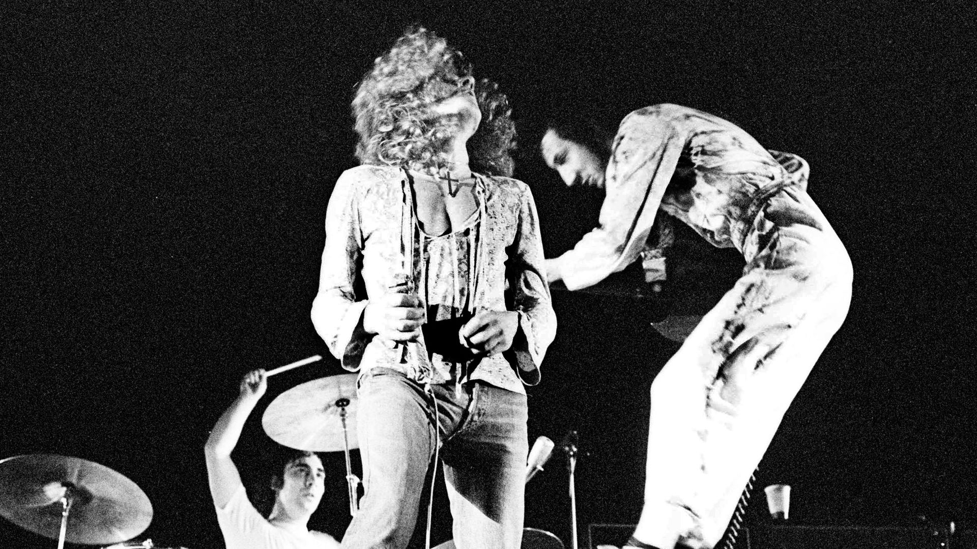 Black-and-white concert scene: a curly-haired lead singer in an open jacket stands center stage with a microphone, guitarist to the right, drummer visible in the back.