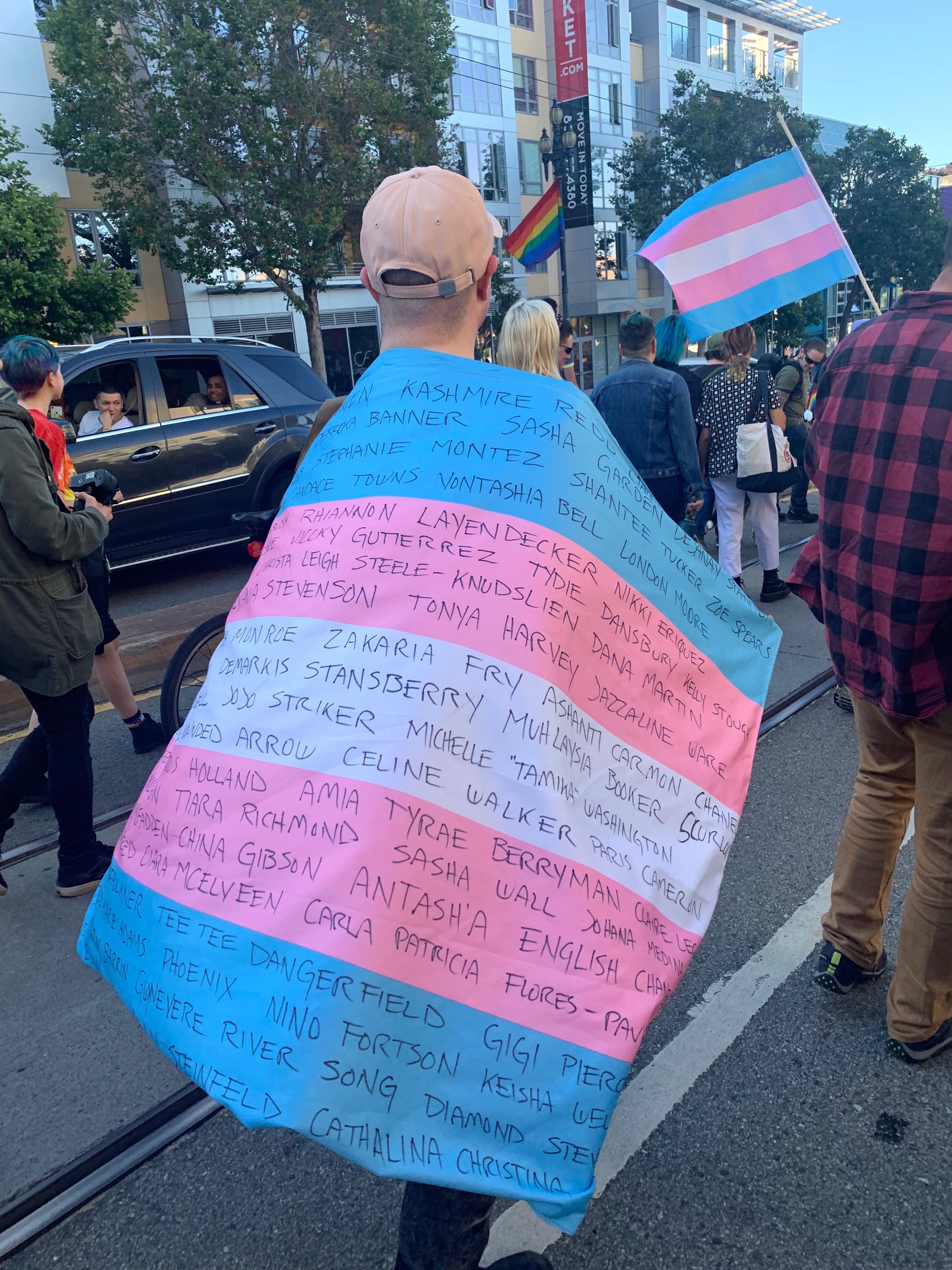 A marcher at San Francisco Pride wearing a trans flag with the names of transgender people who have been killed