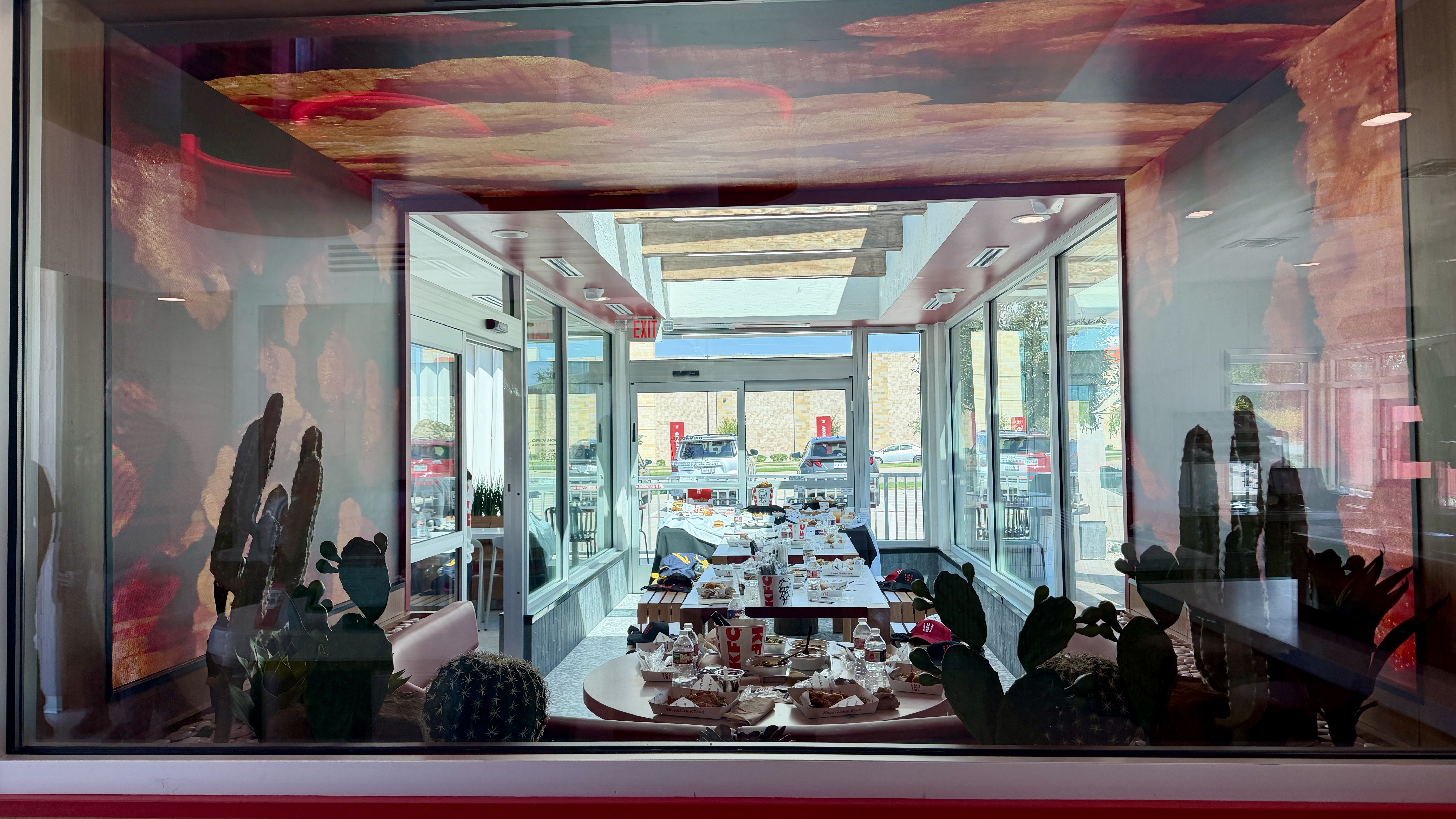 View through a glass window of a bright cafe: a long table piled with KFC takeout boxes, chairs, and potted cacti; skylights with wooden beams, an EXIT sign, and cars outside.