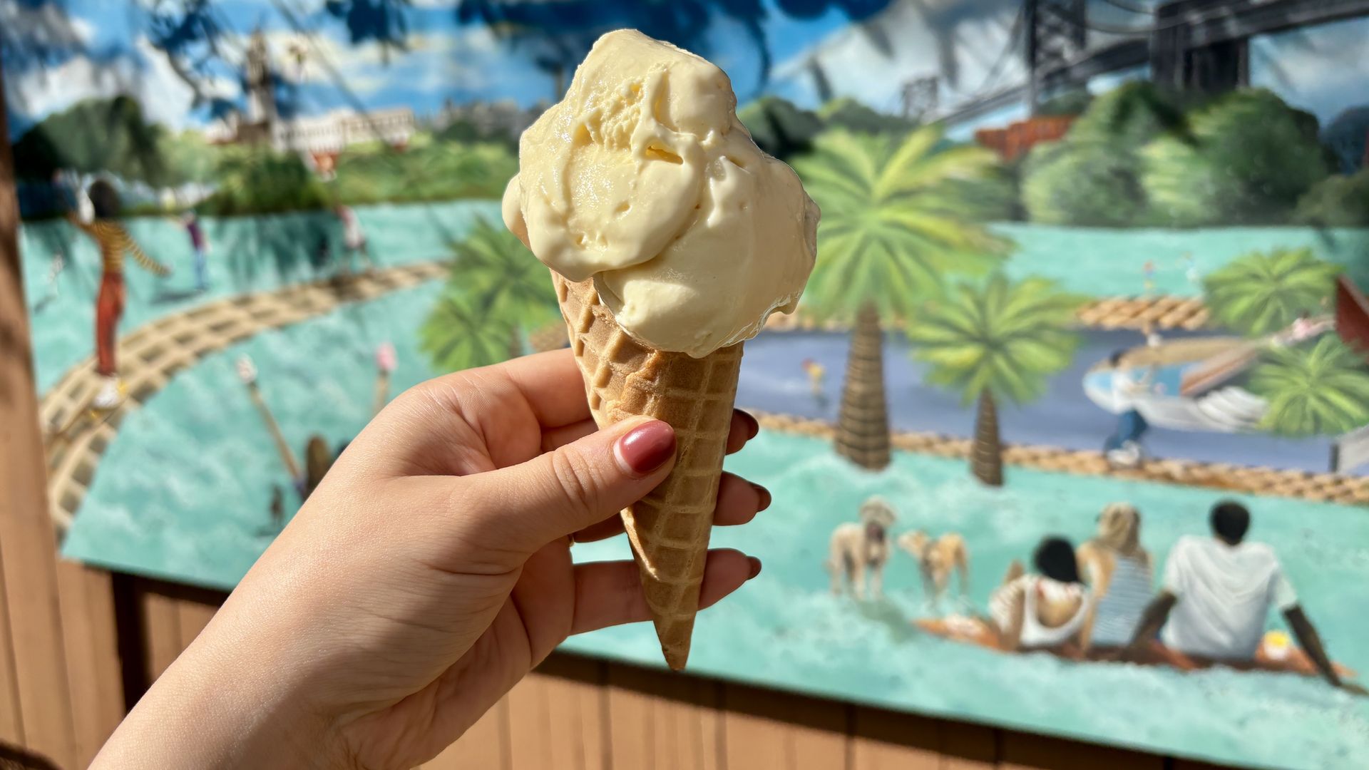 Hand with red nail polish holds a waffle cone topped with two scoops of vanilla ice cream, foreground; bright mural of a tropical pool scene with palm trees and people in the background.