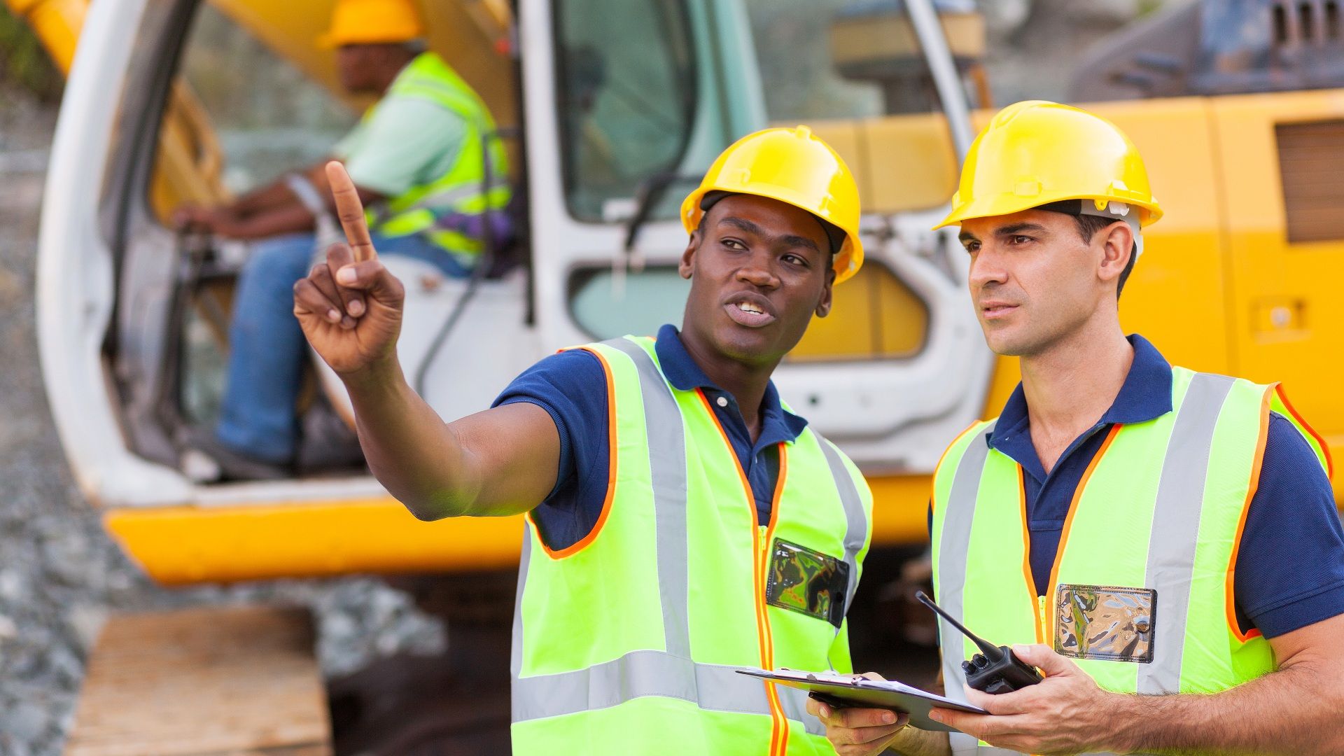 Two construction workers in yellow hard hats and high-visibility vests stand at a construction site; one points and speaks, clipboard in hand, with a yellow excavator in the background.