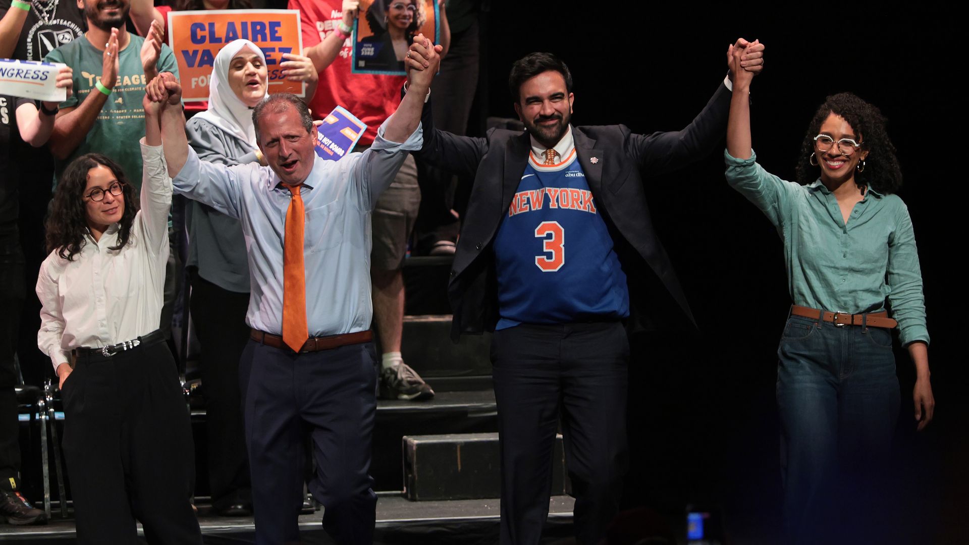 Group on stage raising hands in celebration; foreground shows a man in a light blue shirt with orange tie, and a man in a dark suit with a blue Knicks jersey; a smiling woman in teal with glasses; signs visible in back.
