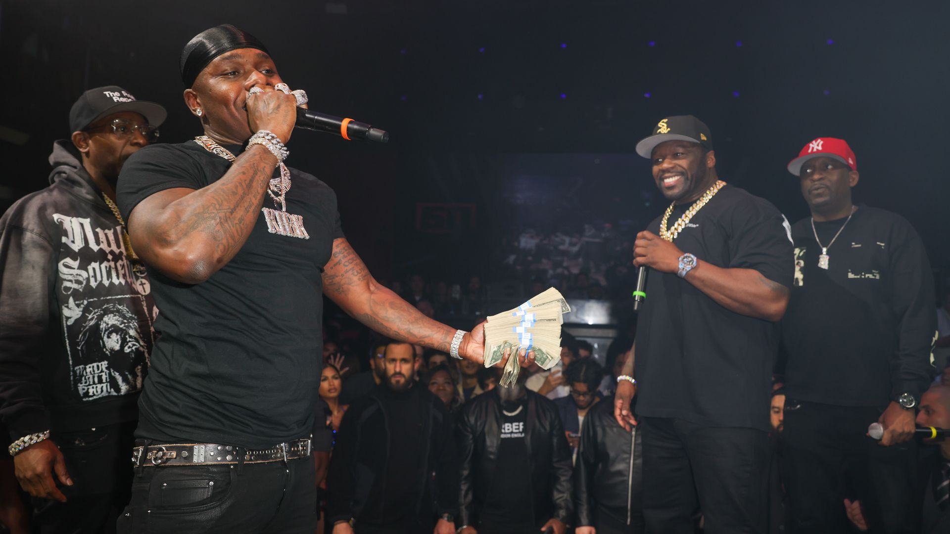 Onstage at a rap concert: a muscular man in a black durag and diamond jewelry raps into a microphone, flashing a stack of cash; two men in black look on as a crowd watches.