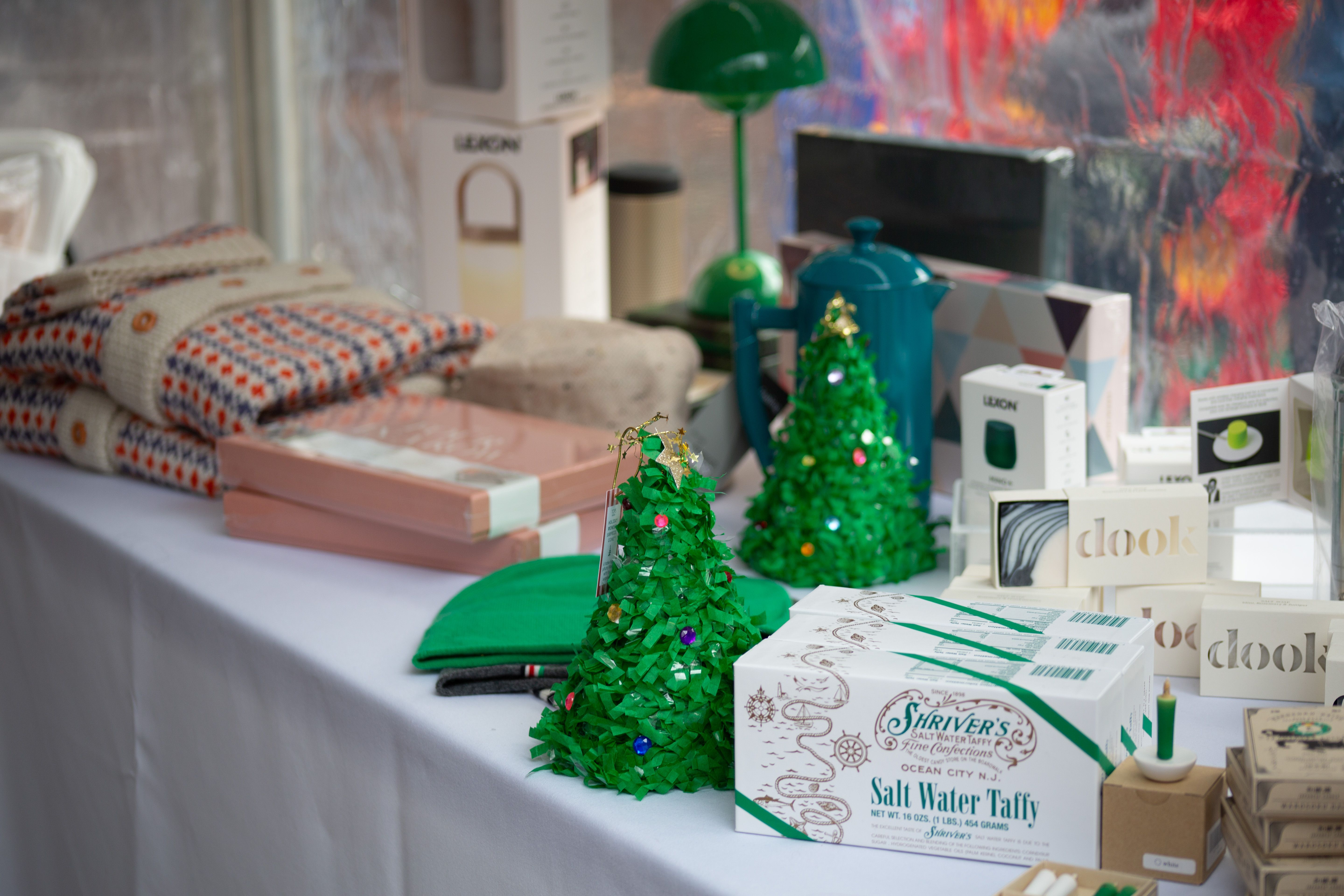 Table display with folded sweaters, green and gray hats, small decorated green Christmas trees, boxes of Shriver's Salt Water Taffy, and various packaged items on a white cloth.