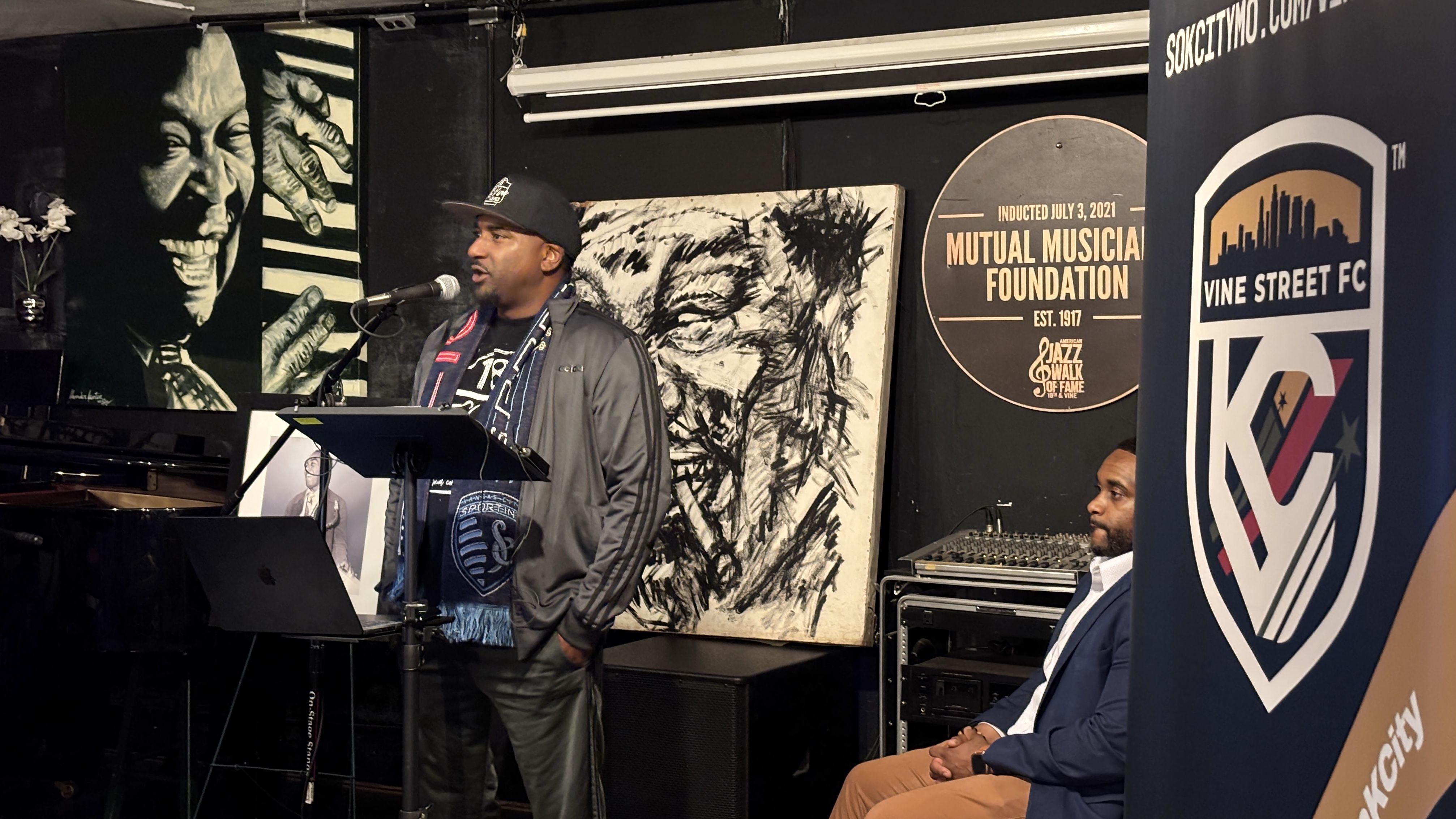 James McGee speaking in a cap and jacket at a microphone on a small stage, with bold black-and-white artwork behind and Dennis Powell seated in a blue blazer to the right; a tall 'VINE STREET FC' banner on the right.