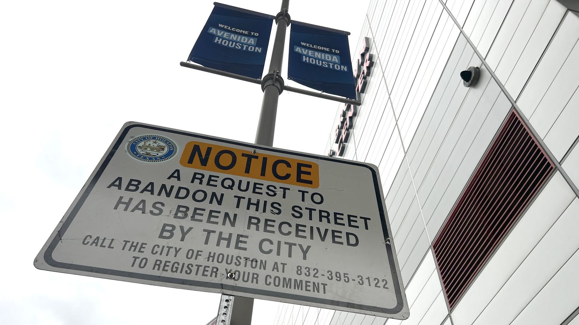 The George R. Brown Convention Center looms over a sign that reads "NOTICE A request to abandon this street has been received by the city. Call the city of Houston at 832-395-3122 to register your comment"