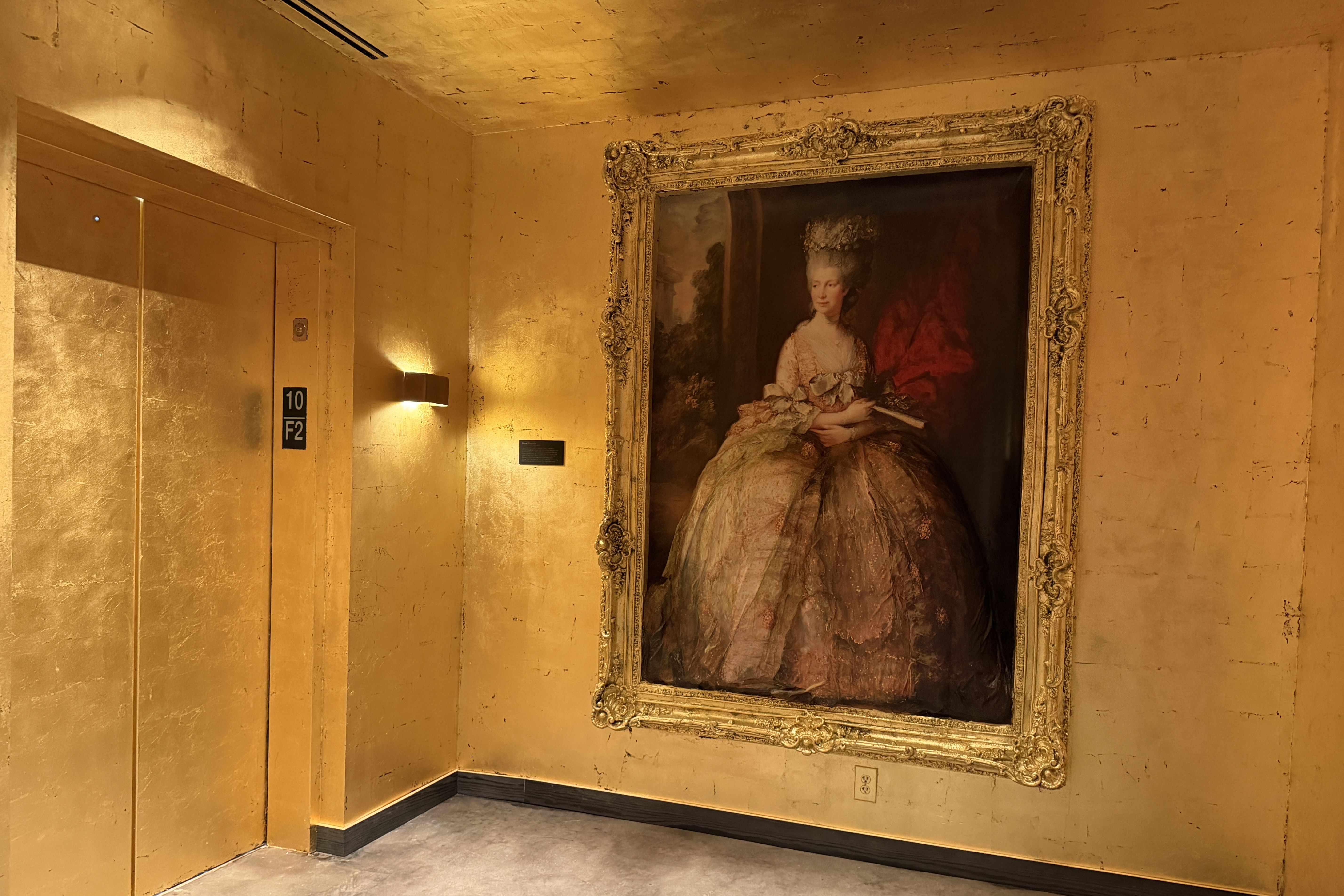 Gold-toned museum corridor featuring a large ornate frame with an 18th-century style portrait of a woman; warm lights illuminate beige walls and a pair of elevator doors labeled 10 F2 on the left.