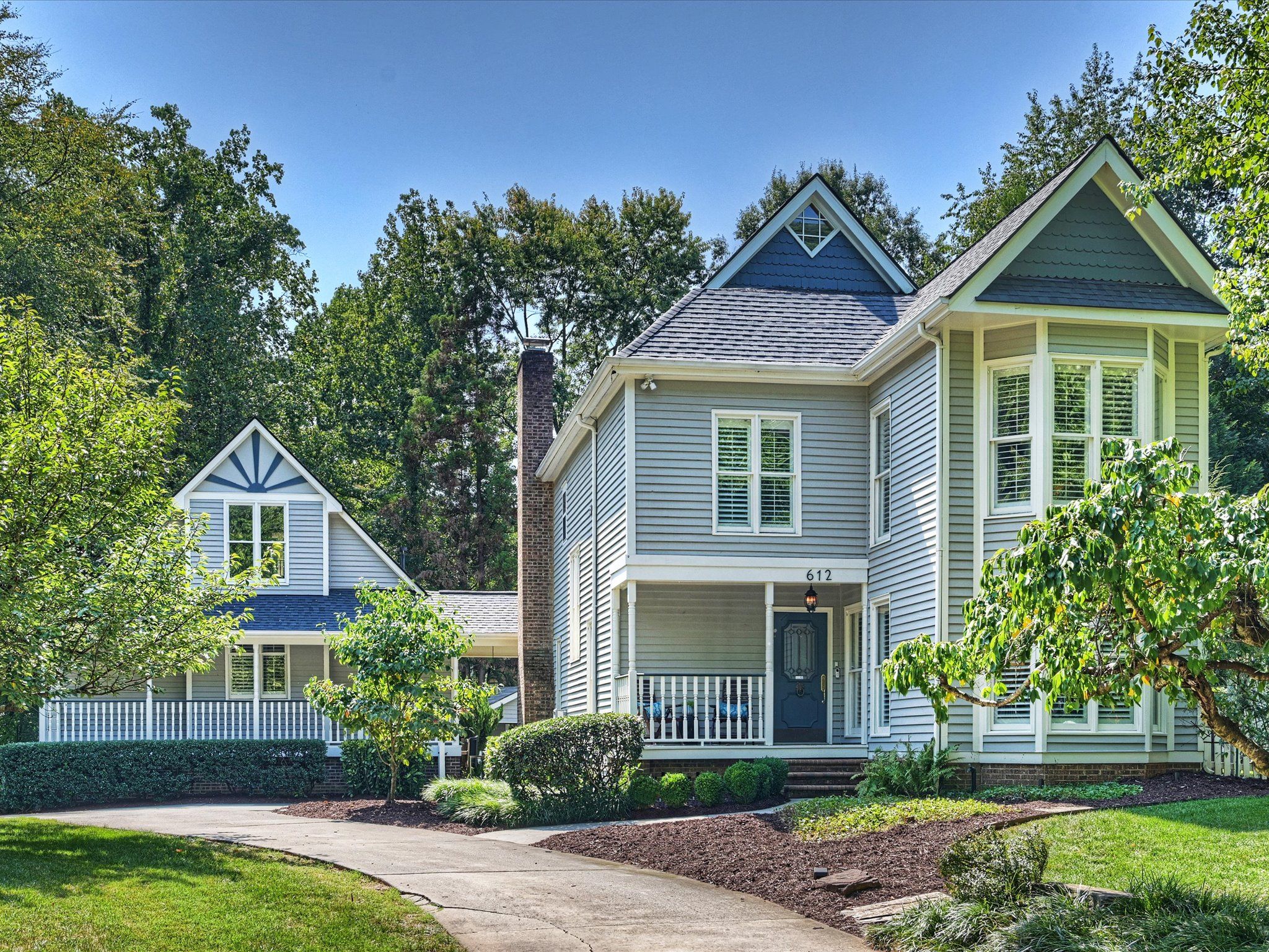 Large two-story gray house with blue and white trim, bay windows, front porch, surrounded by green trees and bushes under a clear blue sky.