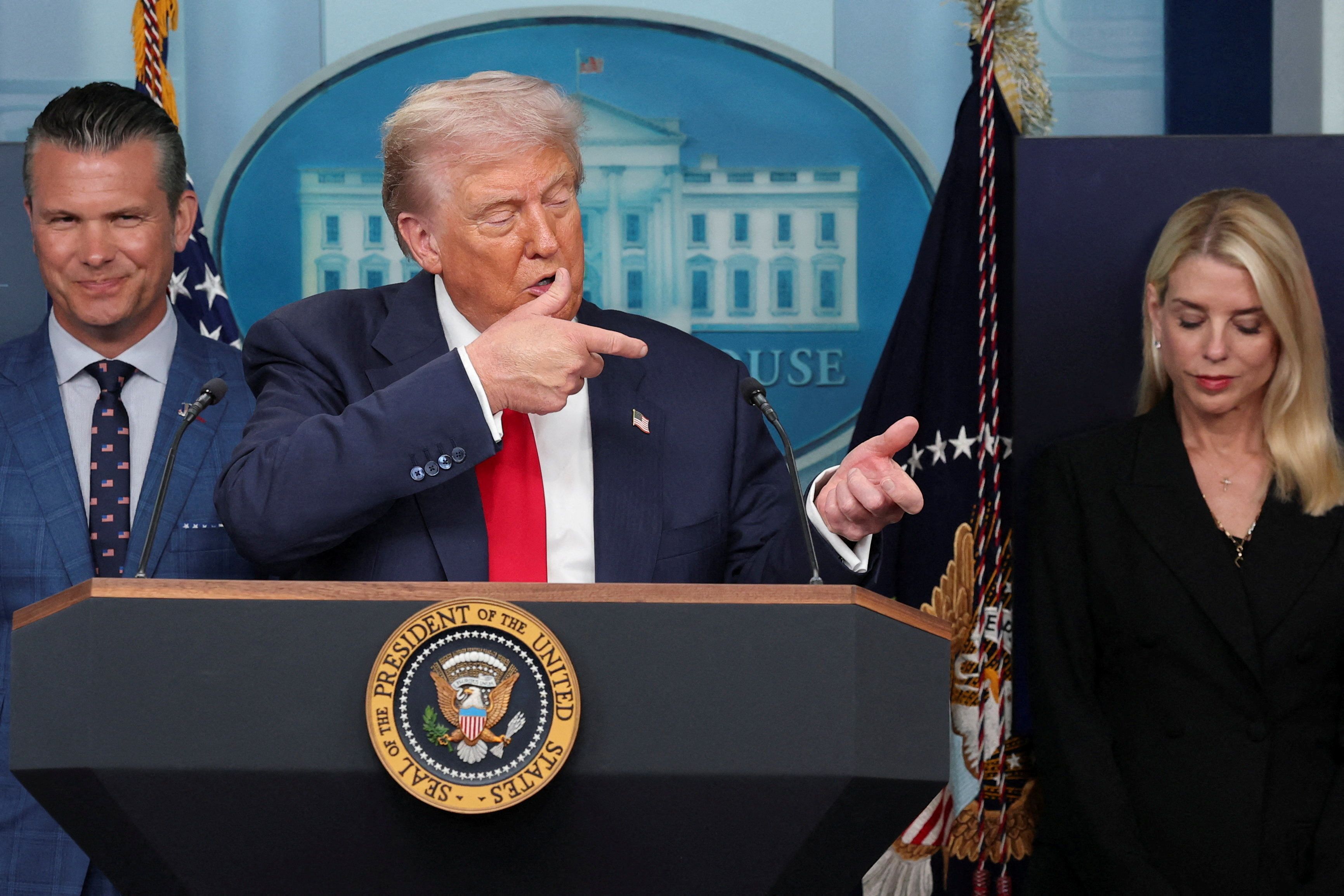 A man with blond hair in a dark suit and red tie stands behind a podium with the presidential seal, gesturing with his hands. Two people stand on either side, one smiling and the other looking down.