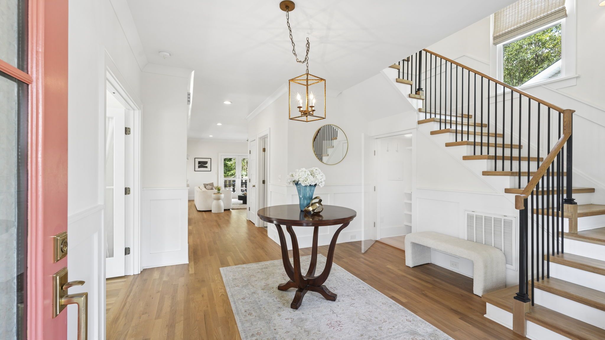 Bright white-entry hallway with hardwood floors. A round dark wood table displays a blue vase of white flowers under a gold geometric chandelier. A black baluster staircase rises to the right; a pink front door sits to the left.