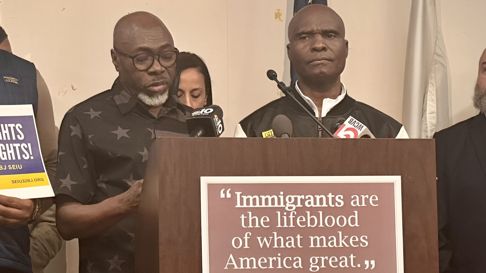 Saint Paul Paul, a Haitian TPS holder who worked at Logan Airport until last year, stands at a lectern and shares how he lost his job alongside a translator (left). 