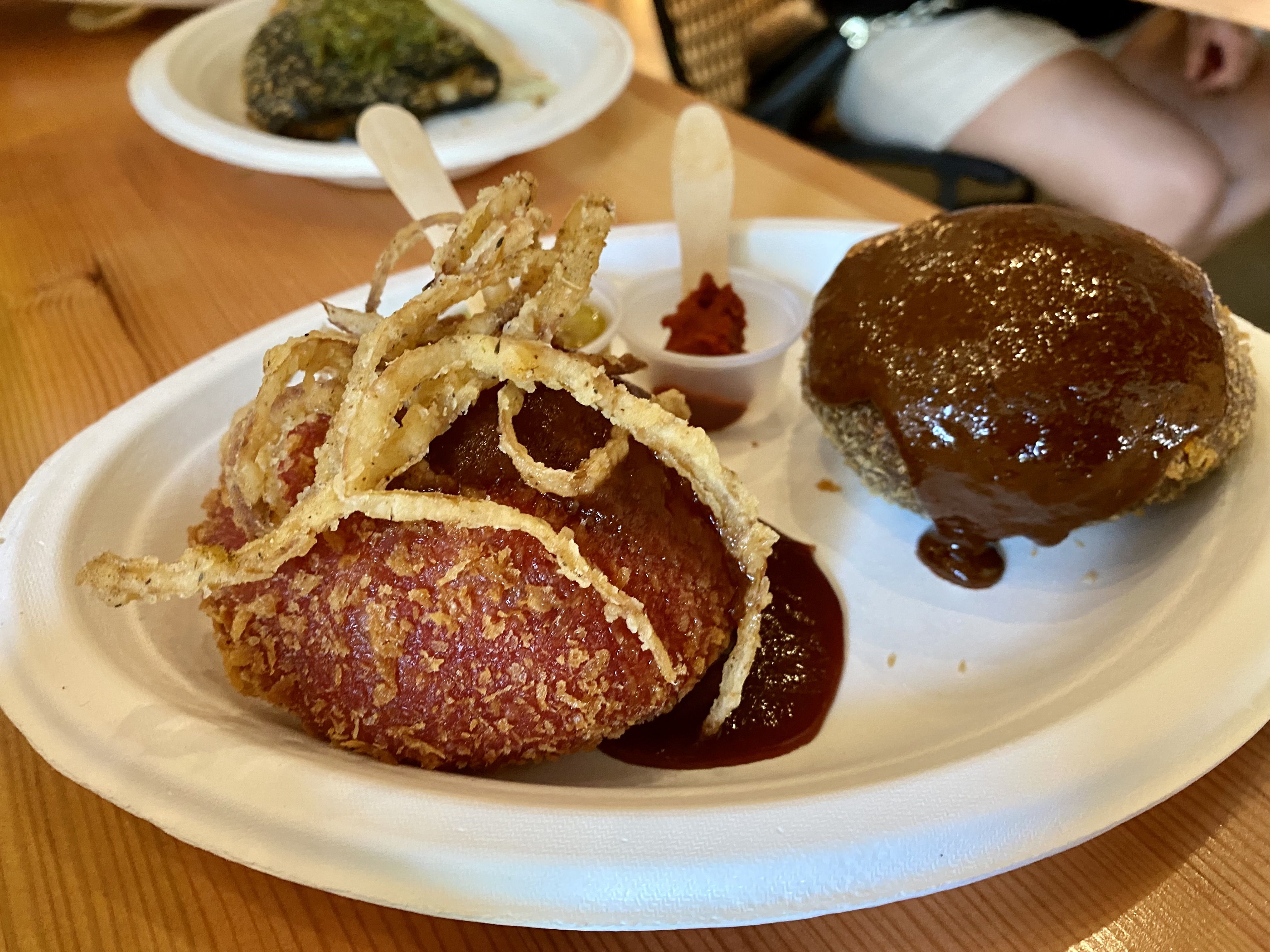 A round reddish colored bun with fried onions on top with another brown pastry behind it. 