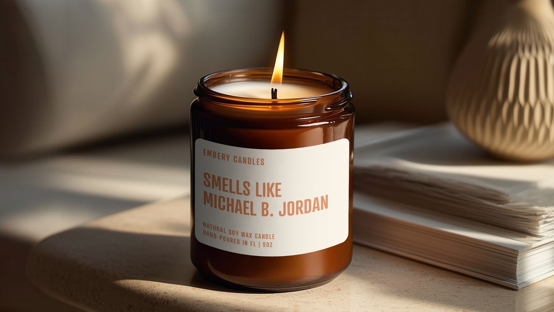 Lit brown candle labeled "Smells Like Michael B. Jordan" on beige table near stacked magazines and beige vase with green plant in soft natural light.