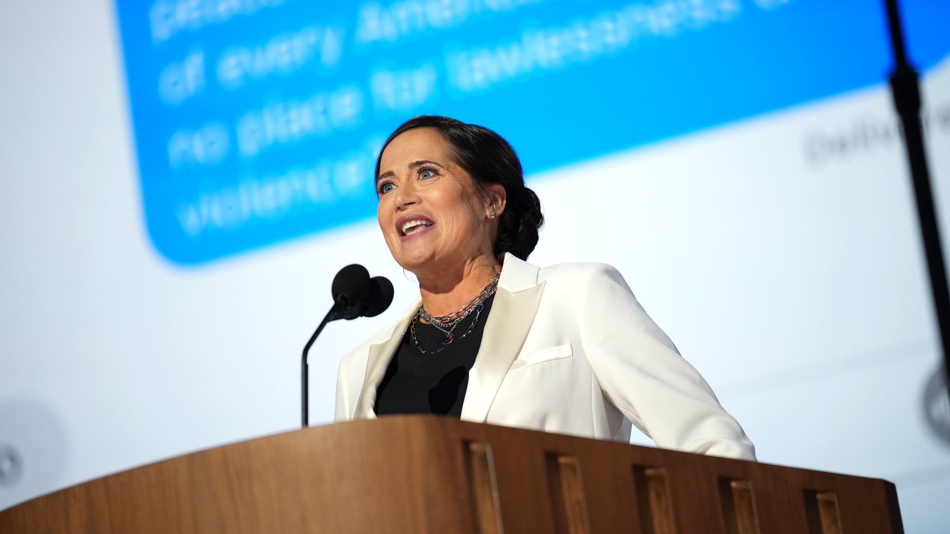 Stephanie Grisham speaks into a microphone in front of a giant screen displaying a text message.