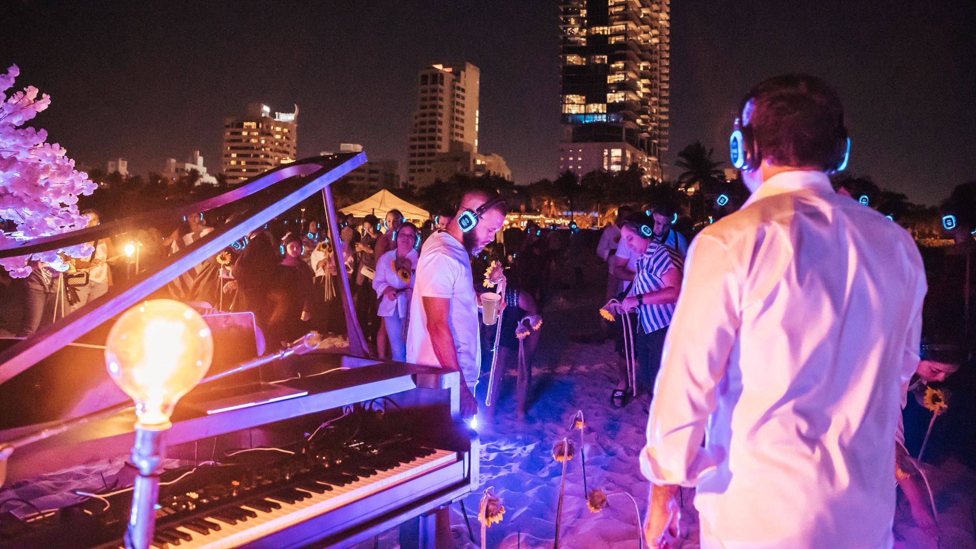 A piano and several people in white shirts wearing over-the-ear headphones lit up by purplish light.