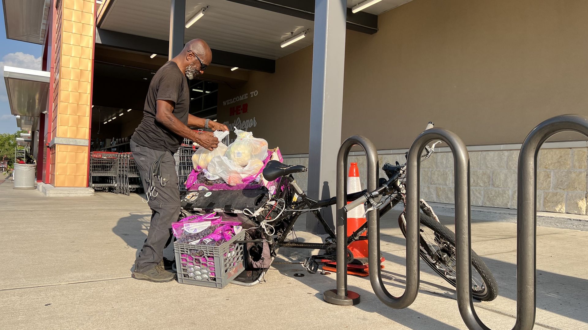 Veon McReynolds loads groceries onto a black bicycle parked at a bike rack outside a large store.