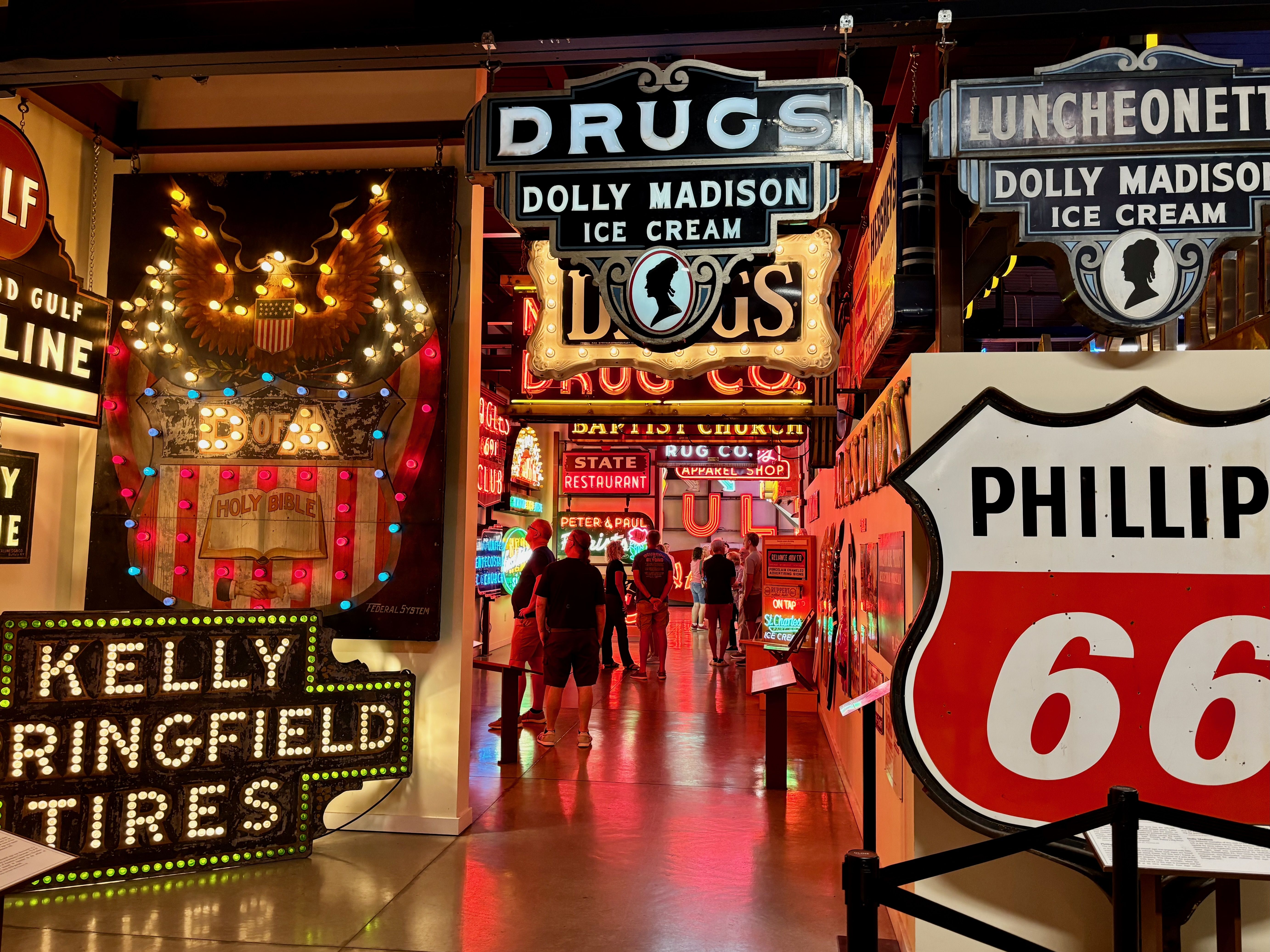 Neon-lit vintage signage scene in a busy arcade or museum, featuring a large "DRUGS" sign and "DOLLY MADISON ICE CREAM" signs, with warm bulbs and visitors walking through.