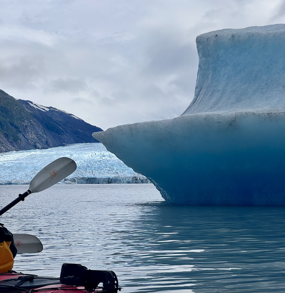 A kayakerβs paddle and gear sit at the edge of calm water beside a massive turquoise-blue iceberg with a curved overhang, while snow-dusted mountains loom in the distant background.