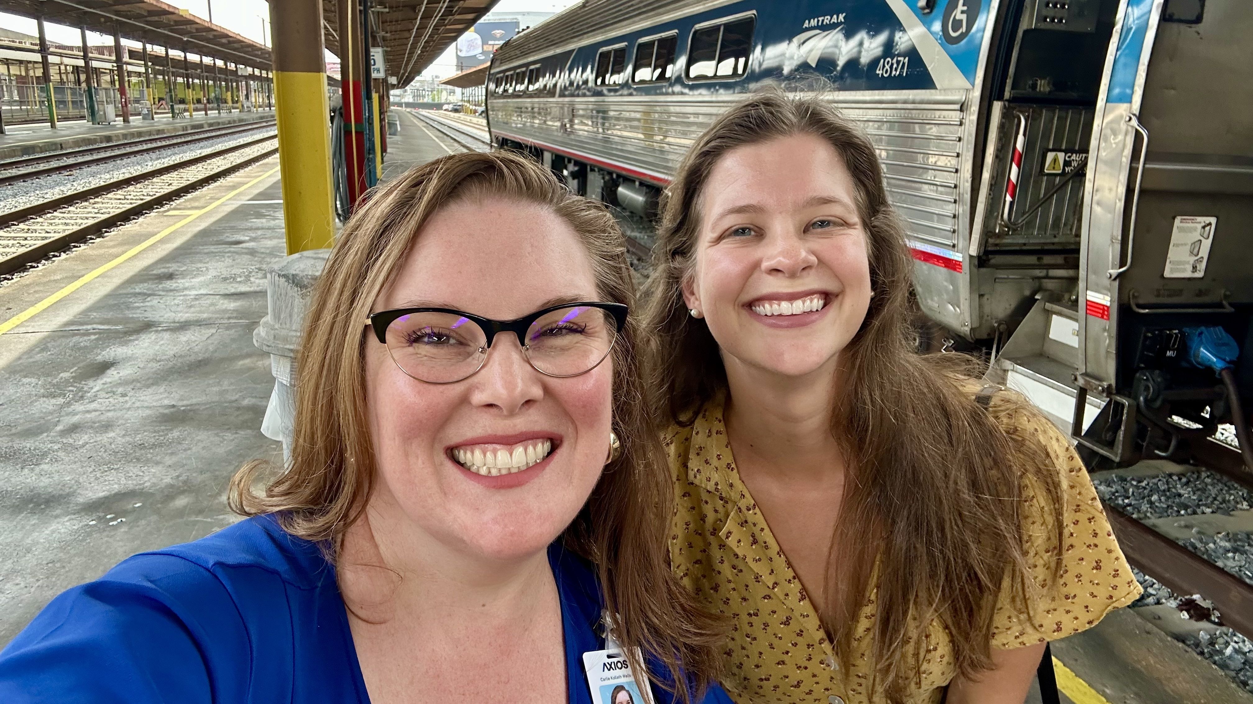 Two smiling women at a train station platform with an Amtrak train behind them. One wears a blue top and glasses, the other a yellow floral dress. Tracks and platform roof visible.