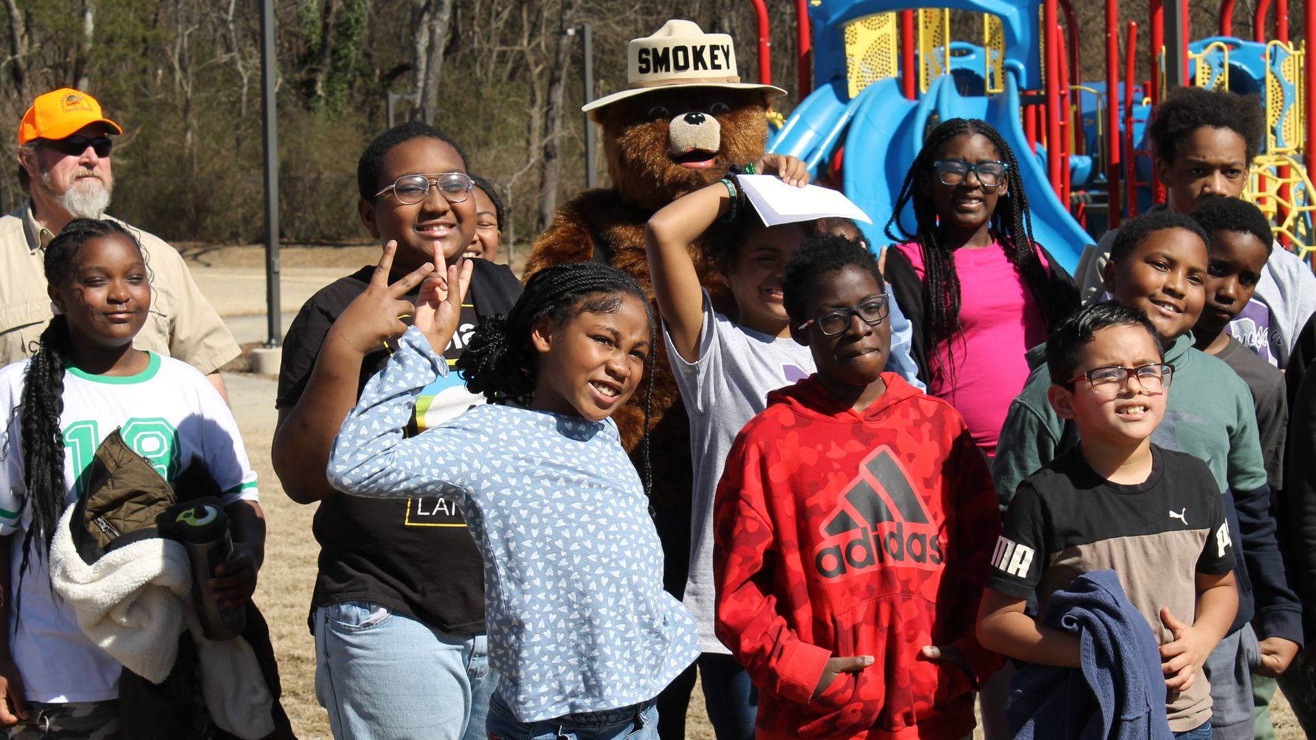 Group of smiling children posing outdoors near blue and red playground equipment with a person dressed as Smokey Bear in the background on a sunny day.