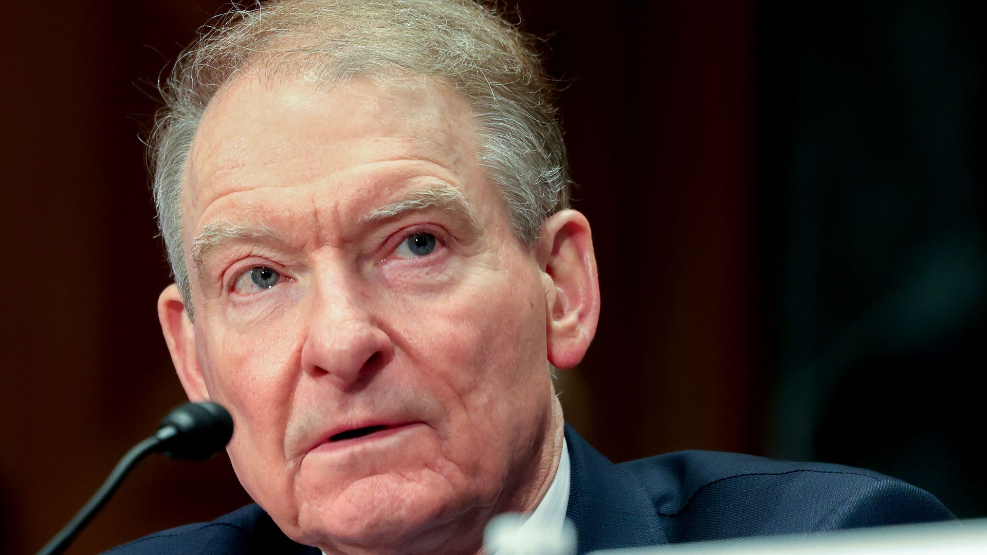 An older man in a suit, speaking on a mic.