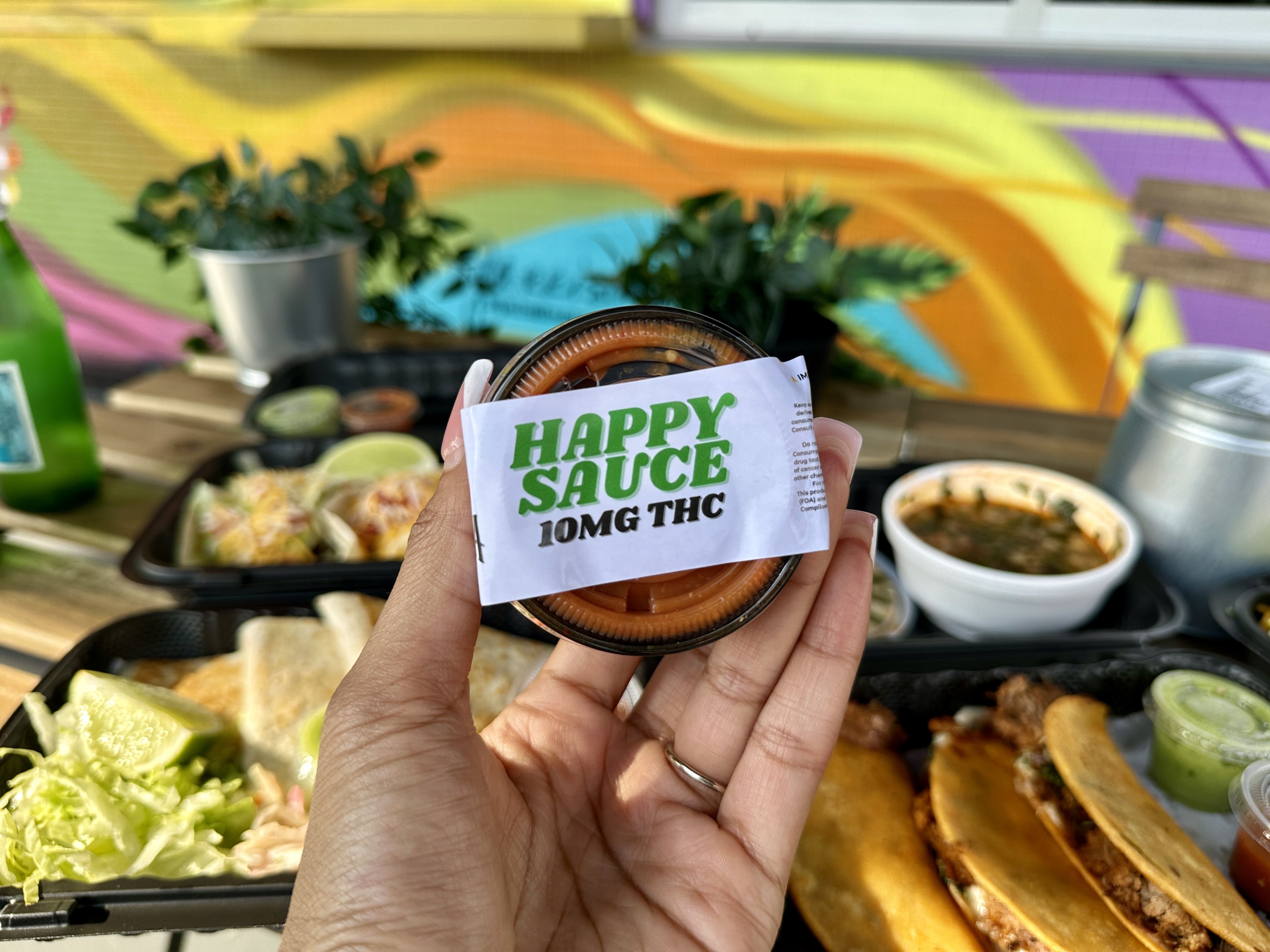 Hand holding a container labeled "Happy Sauce 10MG THC" in front of a table with tacos, lime, lettuce, sauces, and a colorful mural background.