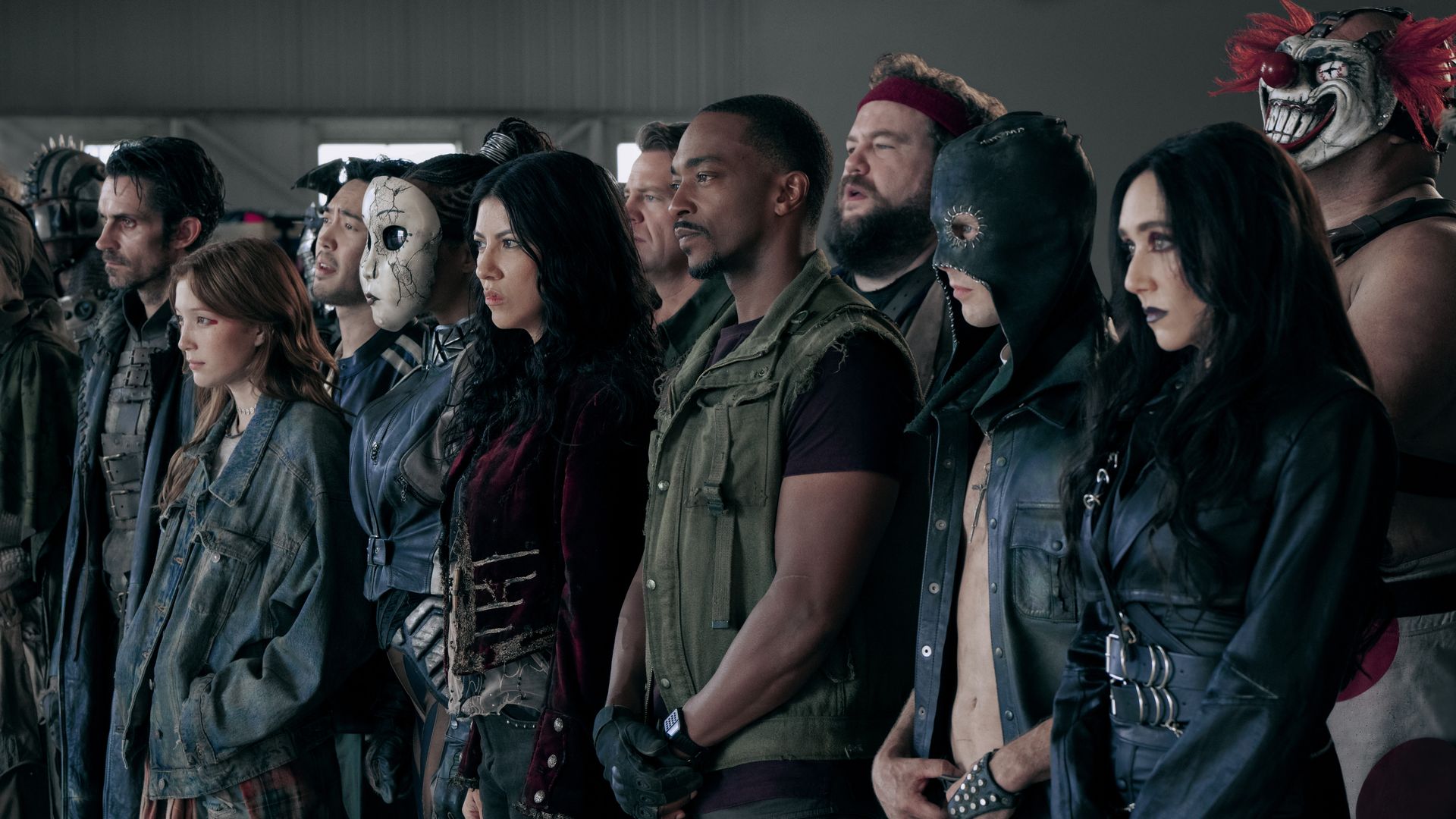 A group of diverse people stand side by side indoors, some in rugged or dark costumes, including a cracked white mask, a black hood, and a clown mask with red hair.